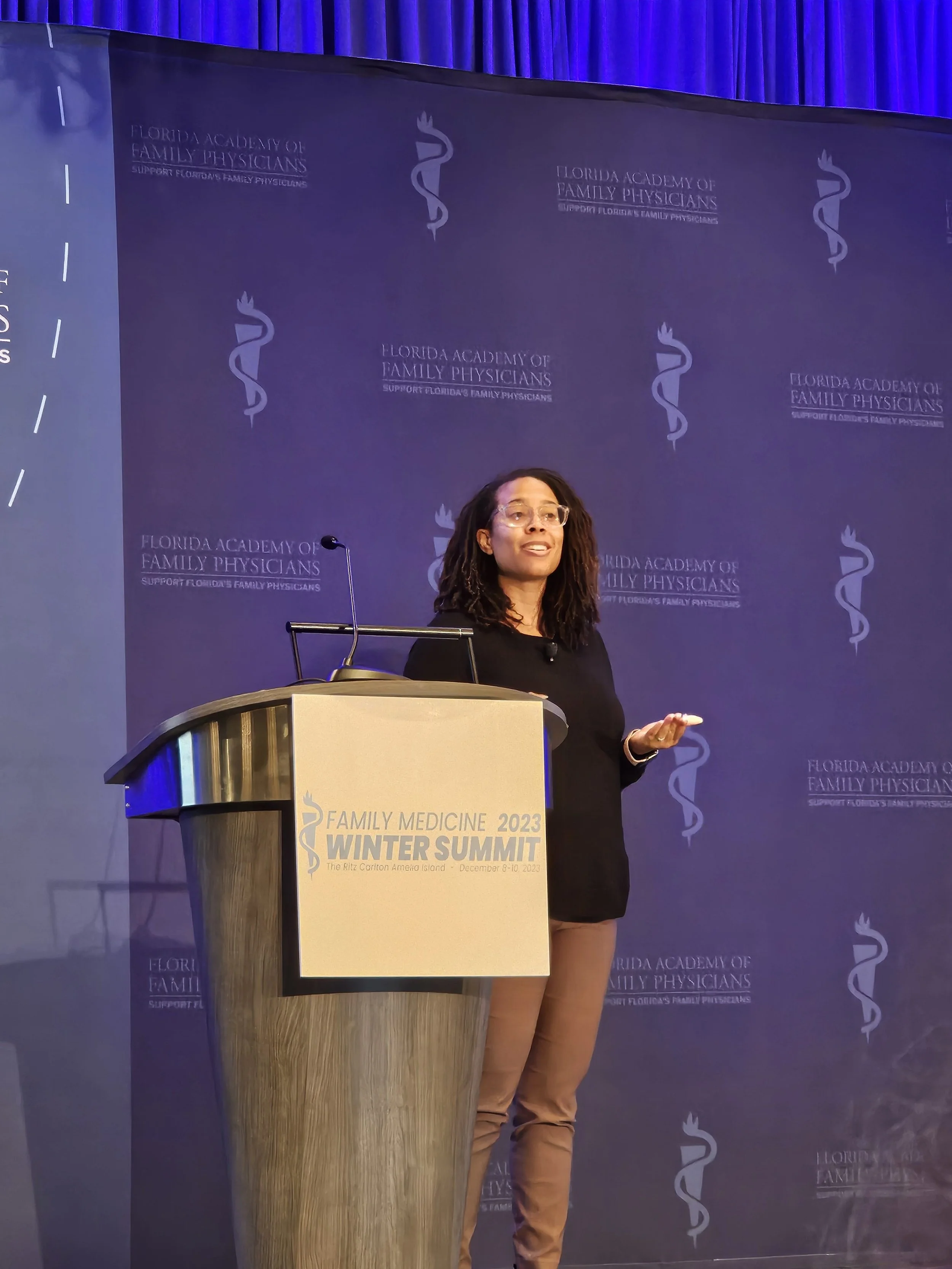 A woman is giving a presentation at the 2023 Winter Summit of the Florida Academy of Family Physicians, standing behind a podium with a sign that says the event details. She is wearing glasses, a black top, and tan pants, and is speaking to the audie