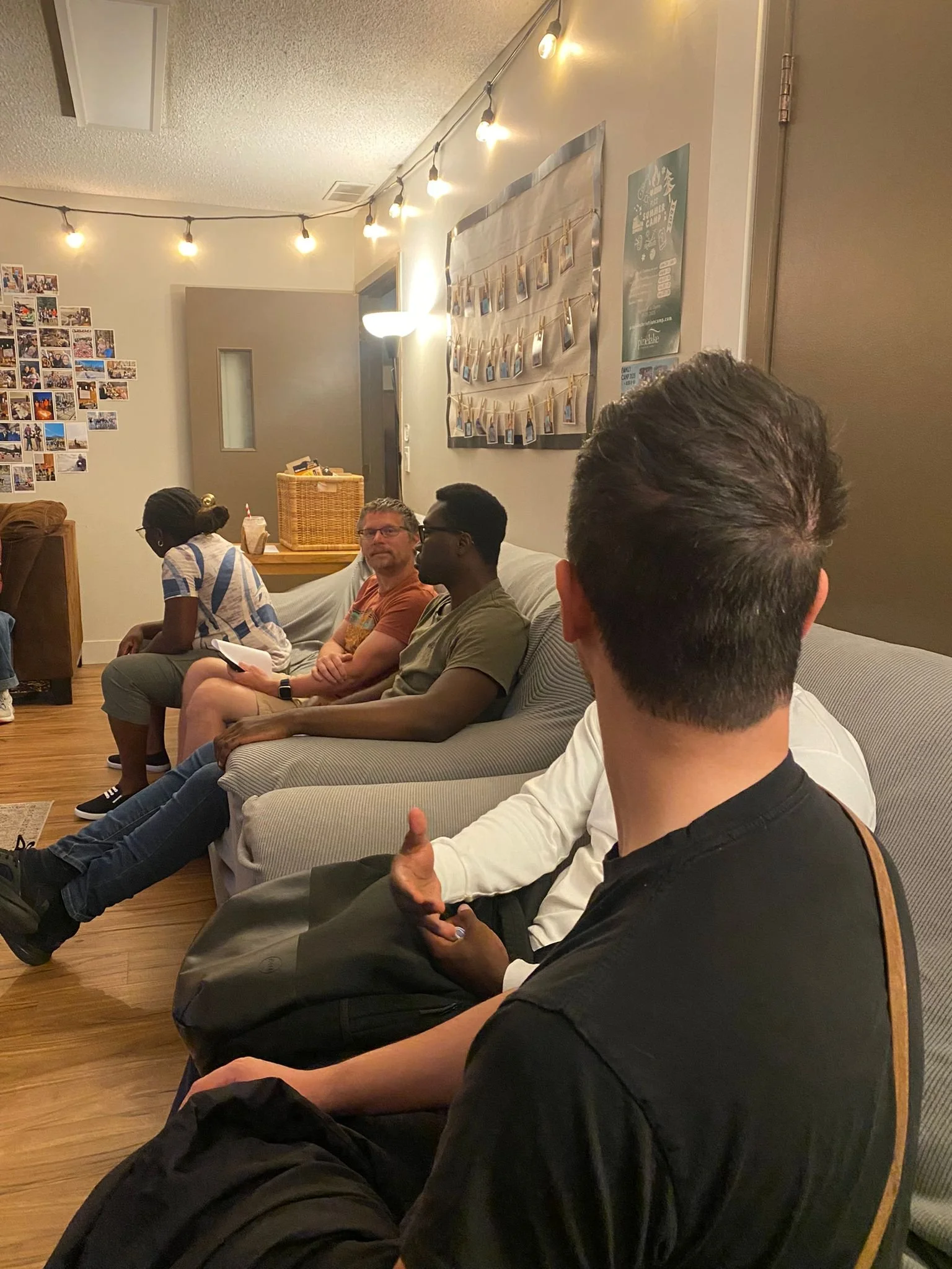 People sitting on a couch in a living room, engaged in conversation. The room has string lights on the ceiling, photo collages, and Polaroids displayed on the walls.