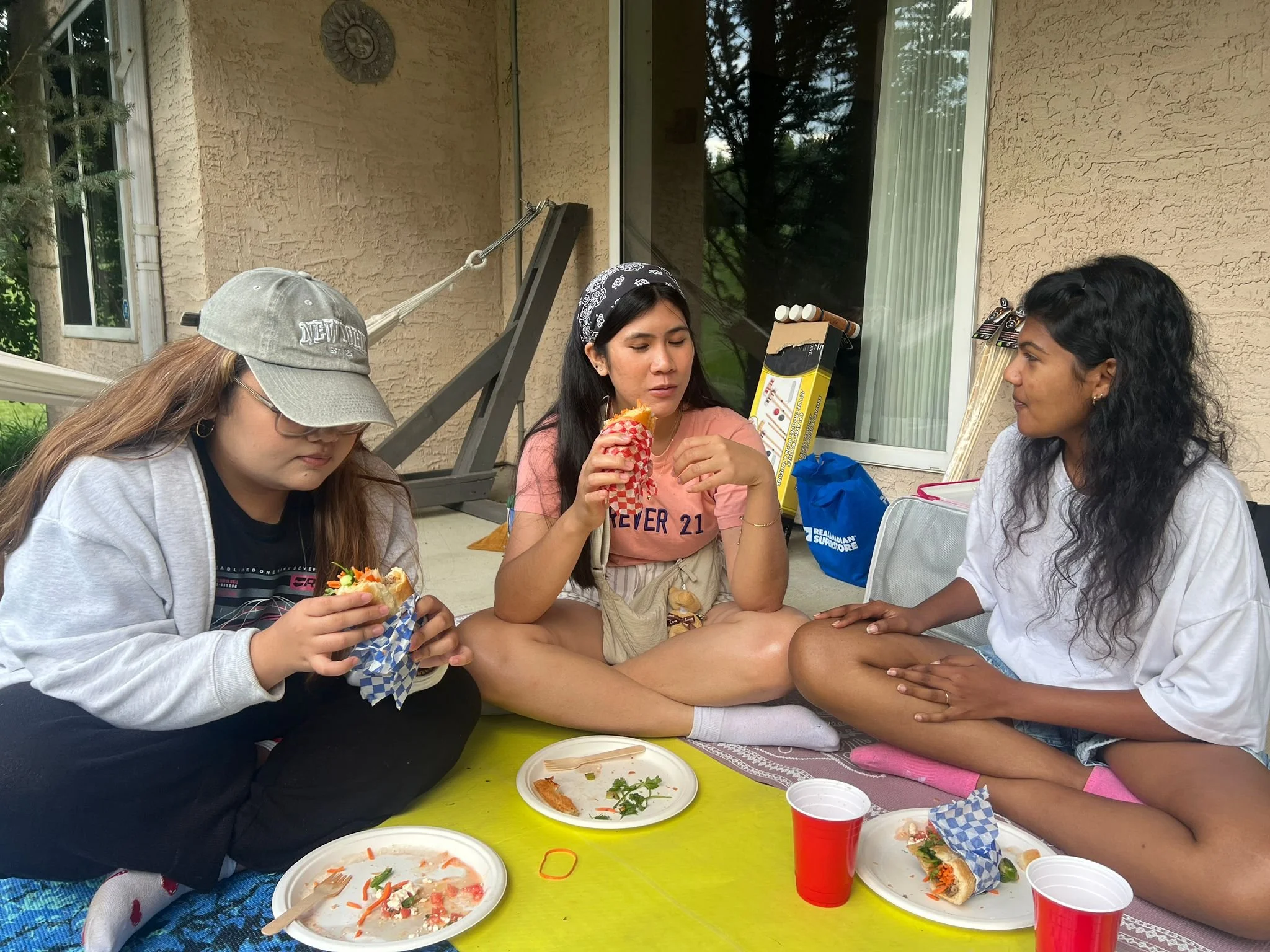 Three women sitting crosses-legged on a yellow table on a porch, eating food. The woman on the left is wearing glasses, a gray cap, a gray hoodie, and a black shirt. The woman in the middle is wearing a bandana, a pink T-shirt, and beige shorts. The woman on the right has curly black hair, is wearing a white T-shirt, and denim shorts. There are dishes with food, red and white cups, and utensils on the table. In the background, there is a window, some blankets, and a box of cooking skewers.