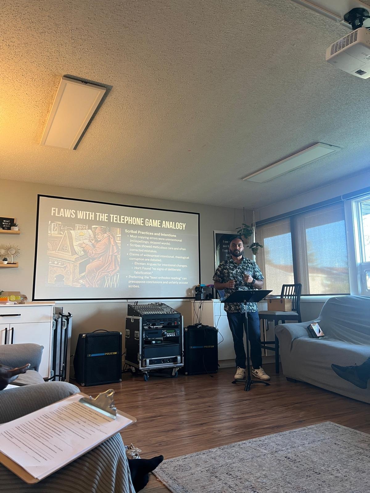 A man stands in front of a presentation screen in a living room, giving a talk on flaws with the telephone game analogy. The room features large windows with blinds, a sofa, and a clipboard held by someone in the foreground.