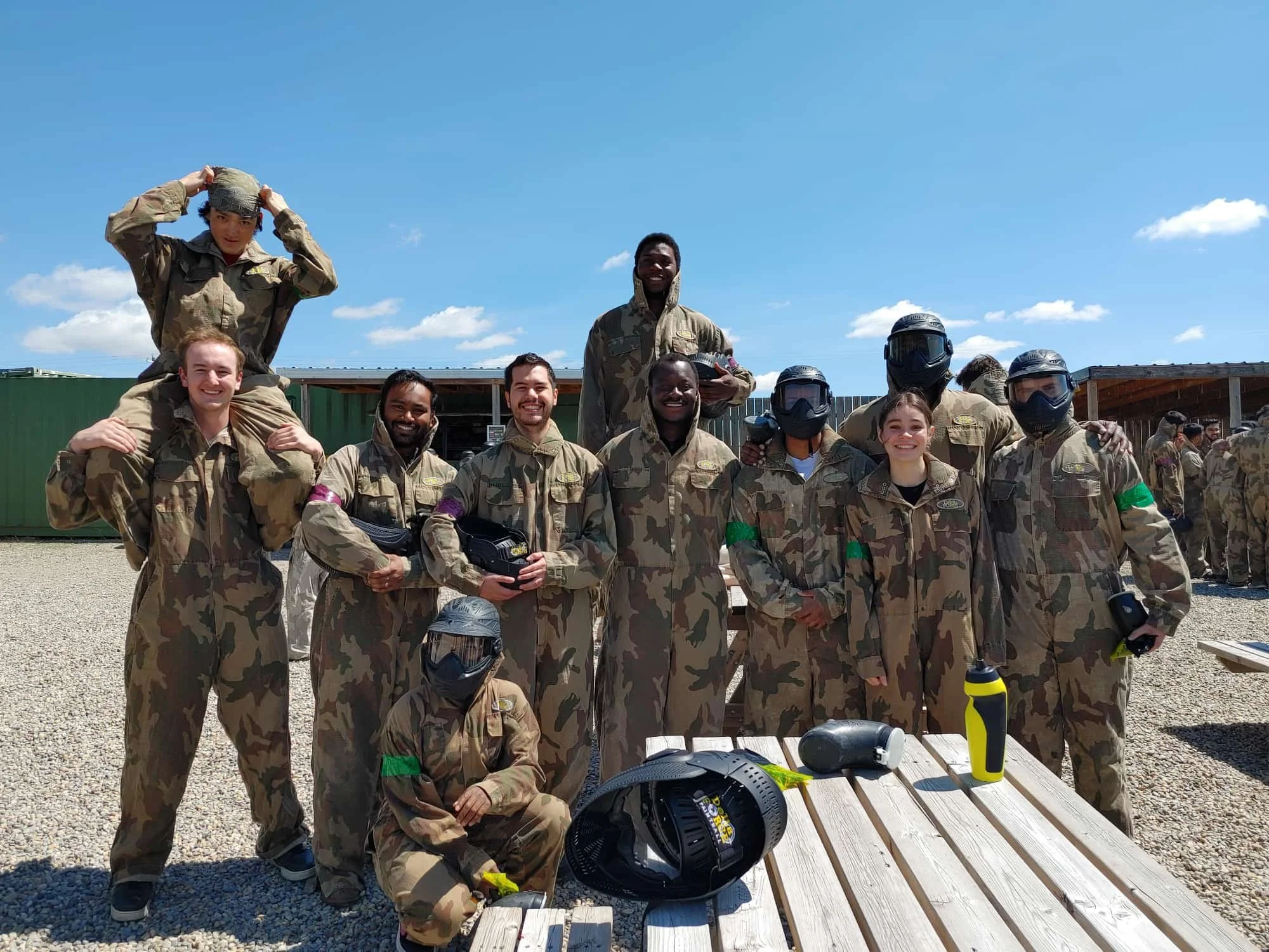 Group of diverse young adults in camouflage military jumpsuits, some wearing helmets, posing outdoors on a sunny day with blue sky and clouds, with tables and other people in similar attire in the background.