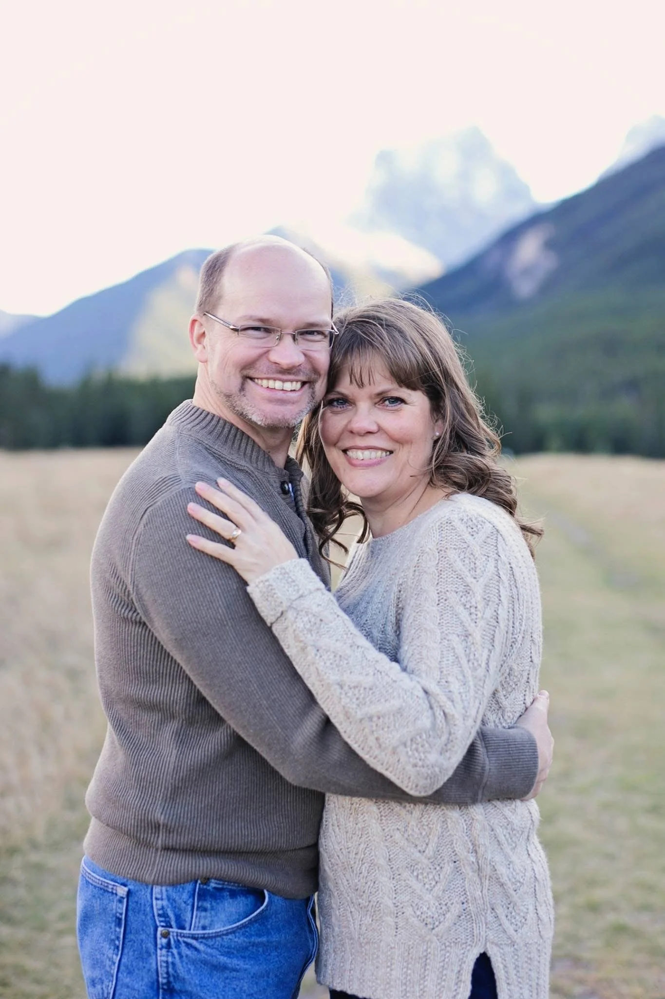 A smiling man and woman hugging outdoors with mountains in the background.
