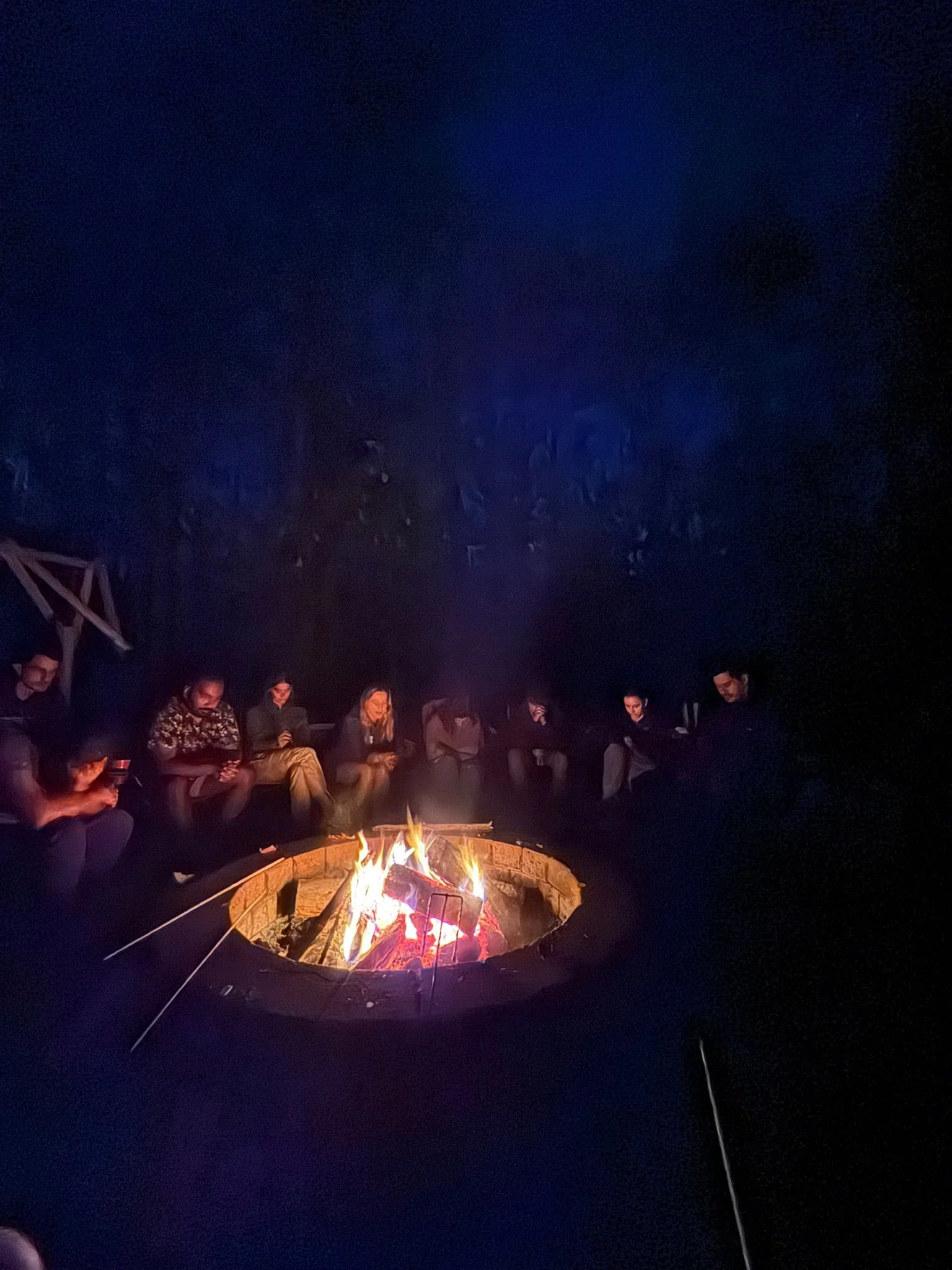 Group of people sitting around a campfire at night in a circle, outdoors.