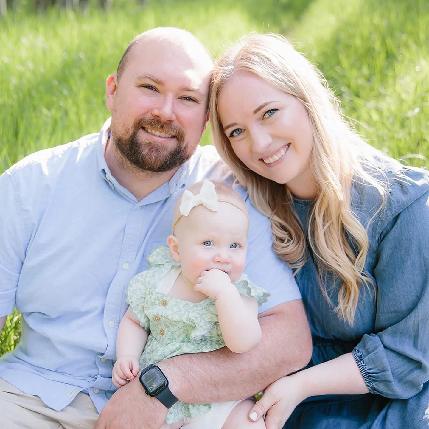 A family of three sitting on grass outdoors, smiling at the camera. The man has a beard and is wearing a light blue shirt, the woman has long blonde hair and is wearing a blue top, and the baby girl has bright blue eyes, a white bow headband, and a green dress.