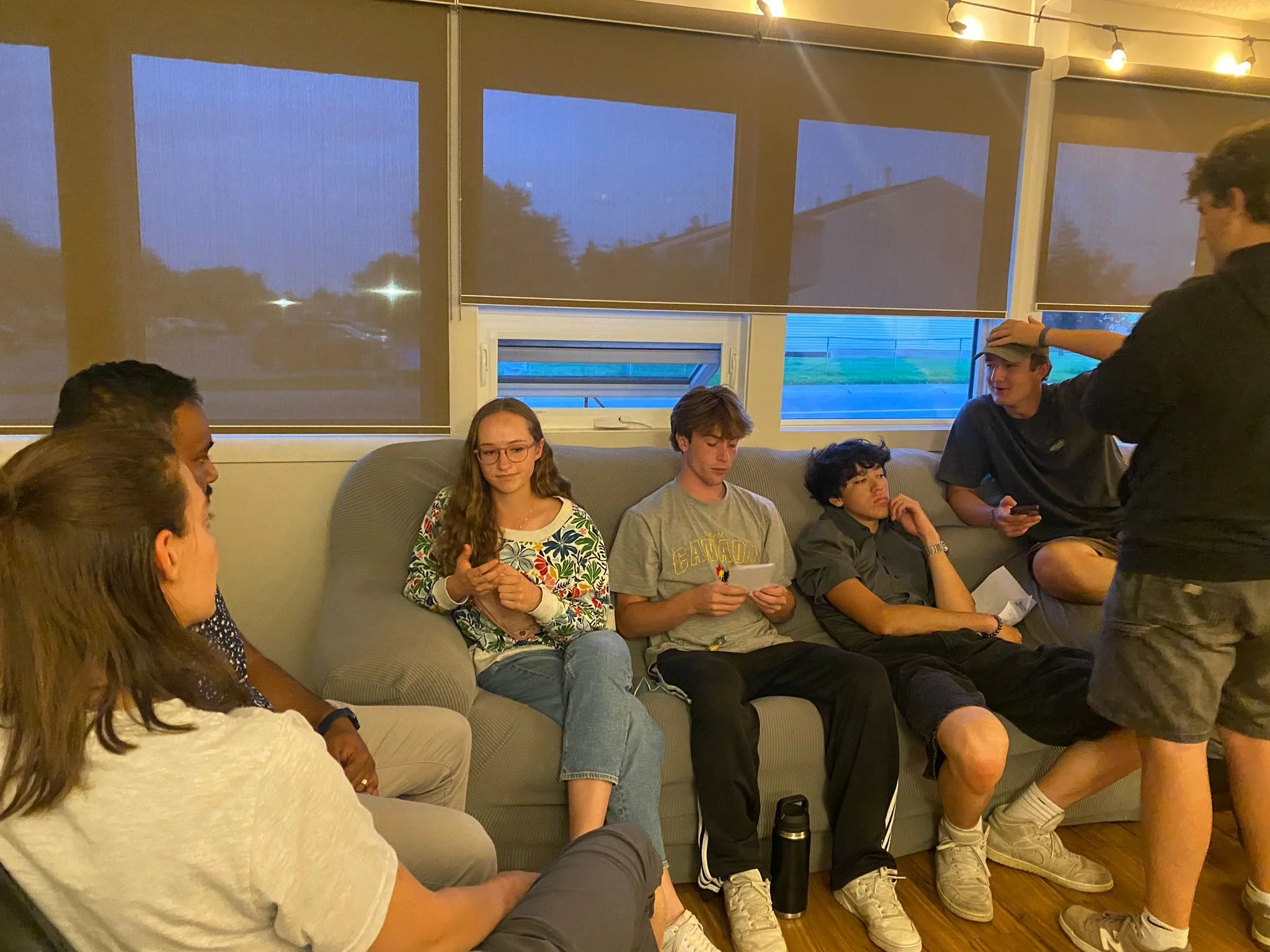 Group of young people sitting on a couch in a living room with large windows and string lights, some engaging with electronic devices.