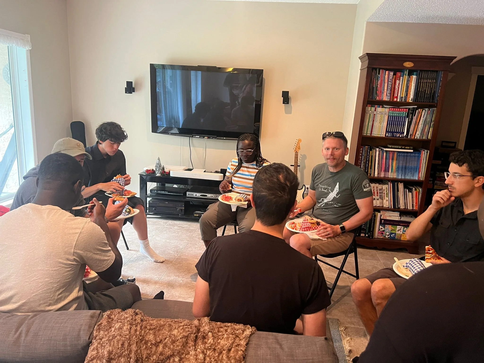 A group of people sitting in a living room, eating pizza, with a large TV on the wall, a bookshelf filled with books, and a guitar in the background.