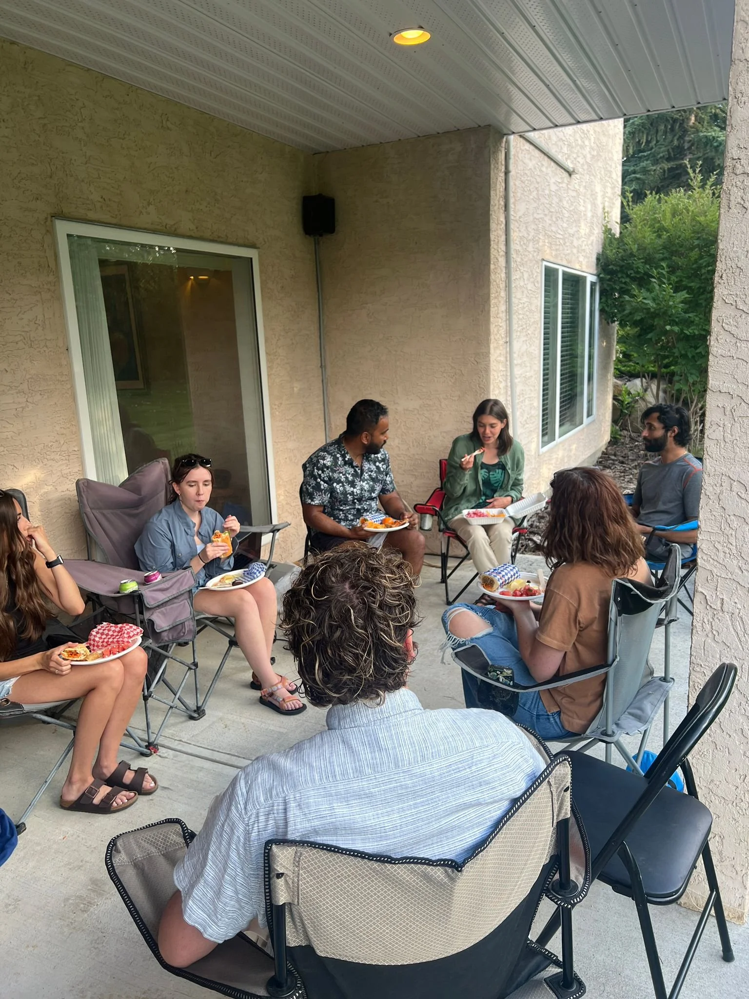 Group of people having a casual outdoor meal on a patio, sitting in chairs with food and drinks, under a roofed area with a beige wall and windows, surrounded by greenery.
