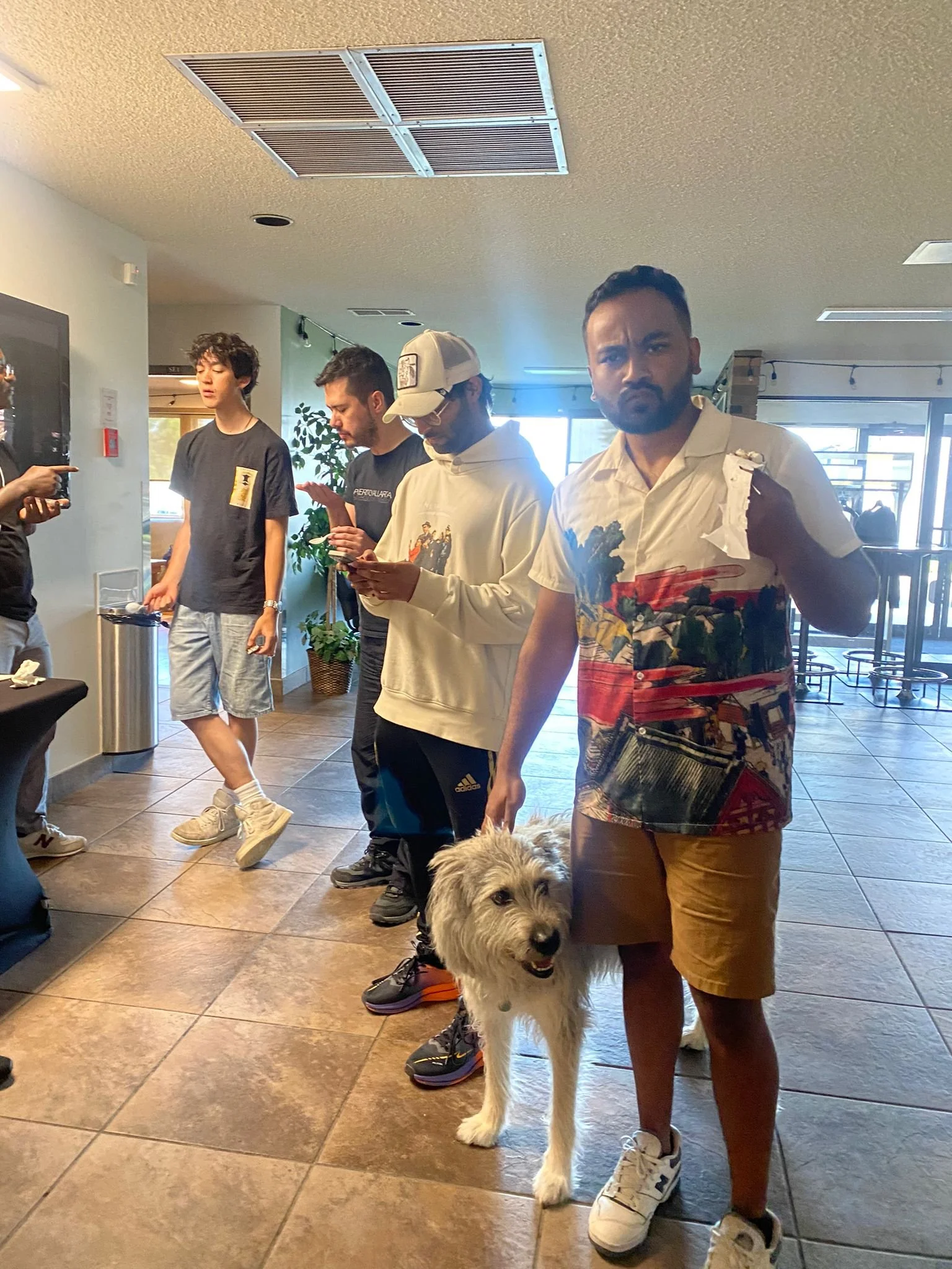 A group of young men standing in a restaurant or cafe with a dog. One man in the foreground is looking at the camera with a serious expression; he is holding the dog on a leash. The others are looking at their phones or talking, and the background shows a tiled floor, plants, and large windows.