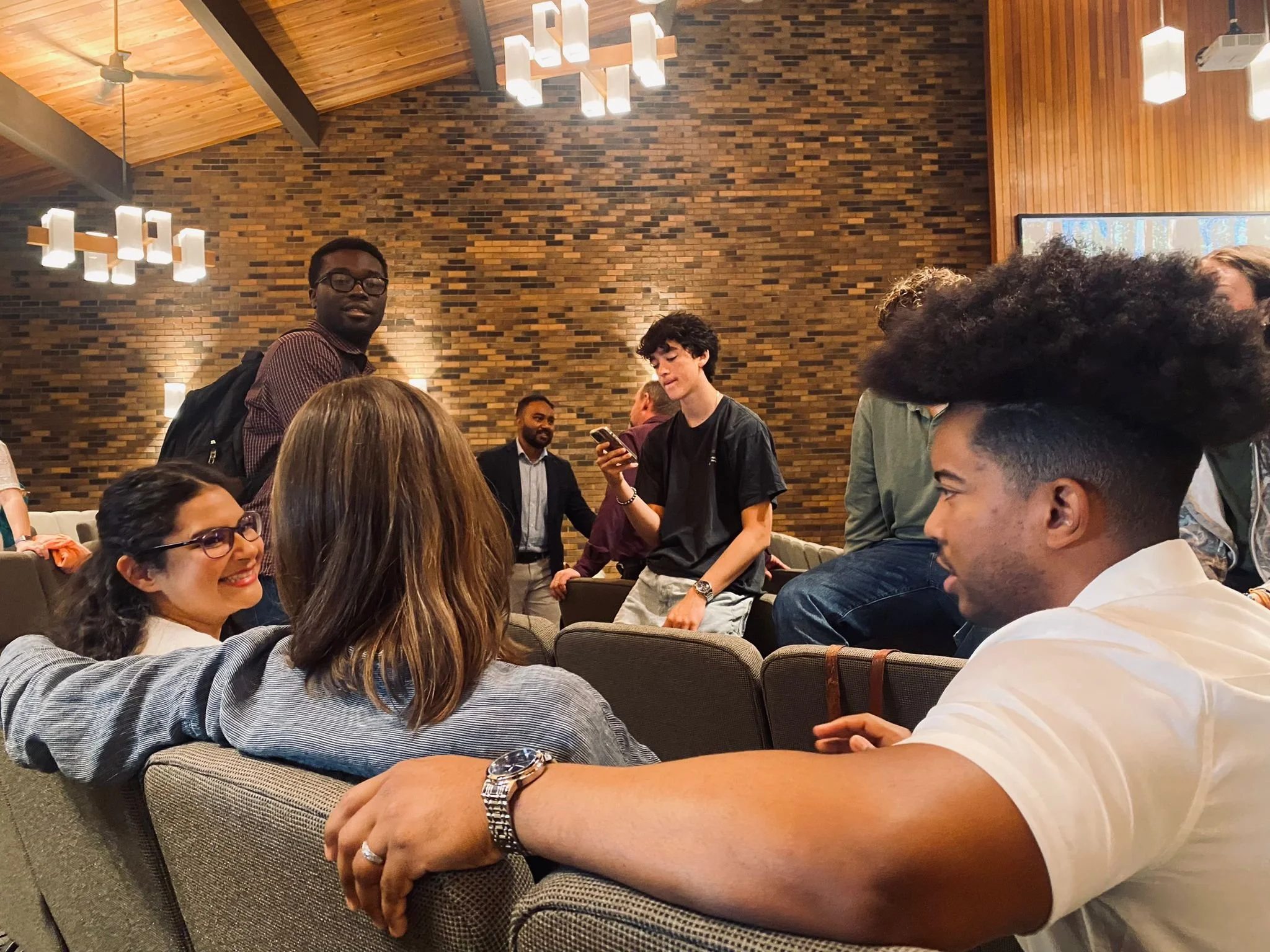 Group of young adults socializing in a warm, wood-paneled room with brick walls, sitting and standing on gray cushioned chairs, with some engaged in conversation and one person looking at his phone.