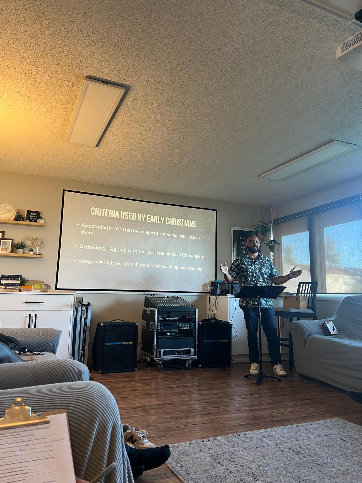 A man giving a presentation in a room with a large window, sofa, and a projection screen displaying criteria used by early Christians.