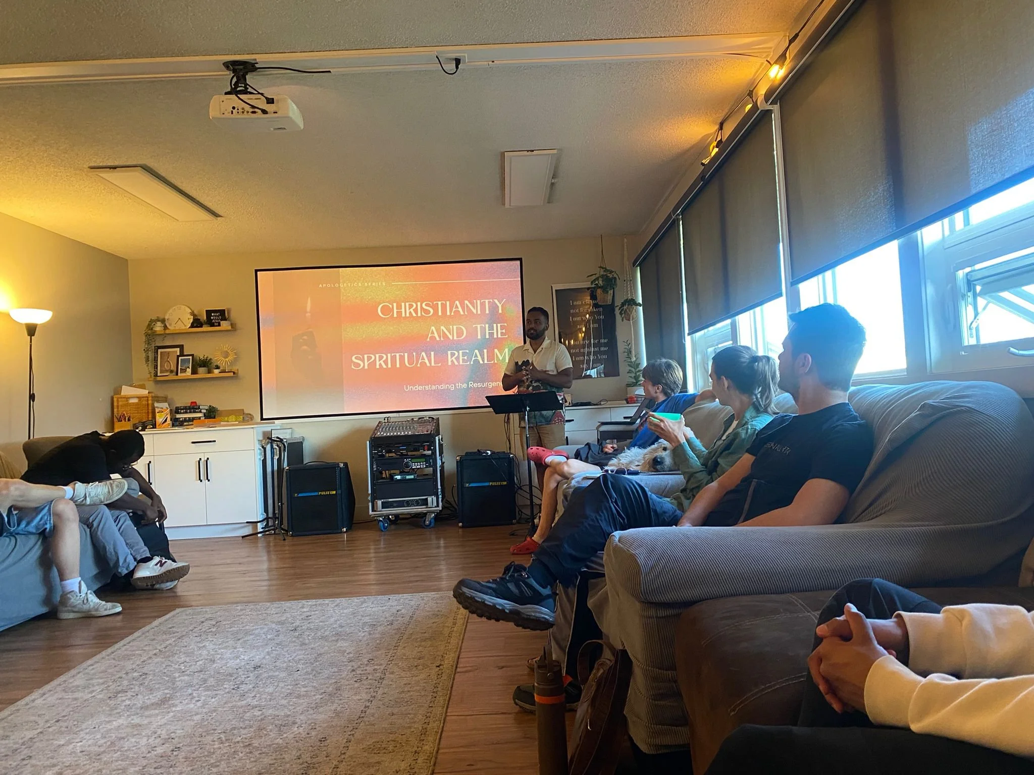 A group of people attending a presentation on Christianity and the spiritual realm in a cozy room with a large screen, facing a speaker standing at a podium. The room has windows along one side, and the audience is seated on a couch and chairs.