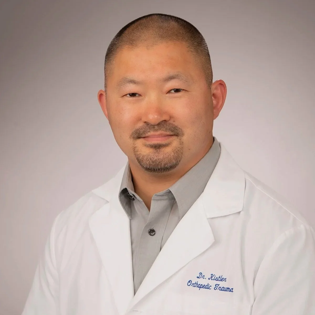 Portrait of Dr. Kistler, an orthopedic trauma doctor, wearing a white coat and gray shirt, with a neutral gray background.