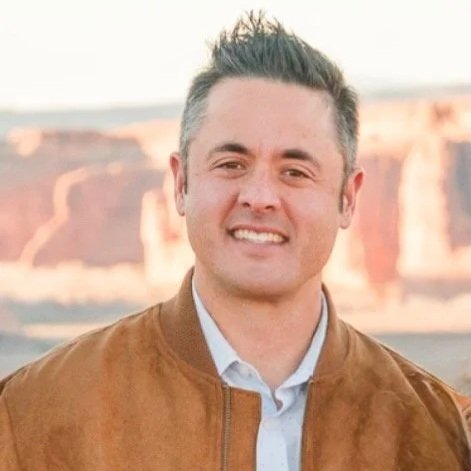 A man with short, dark hair smiling outdoors with a desert landscape in the background.