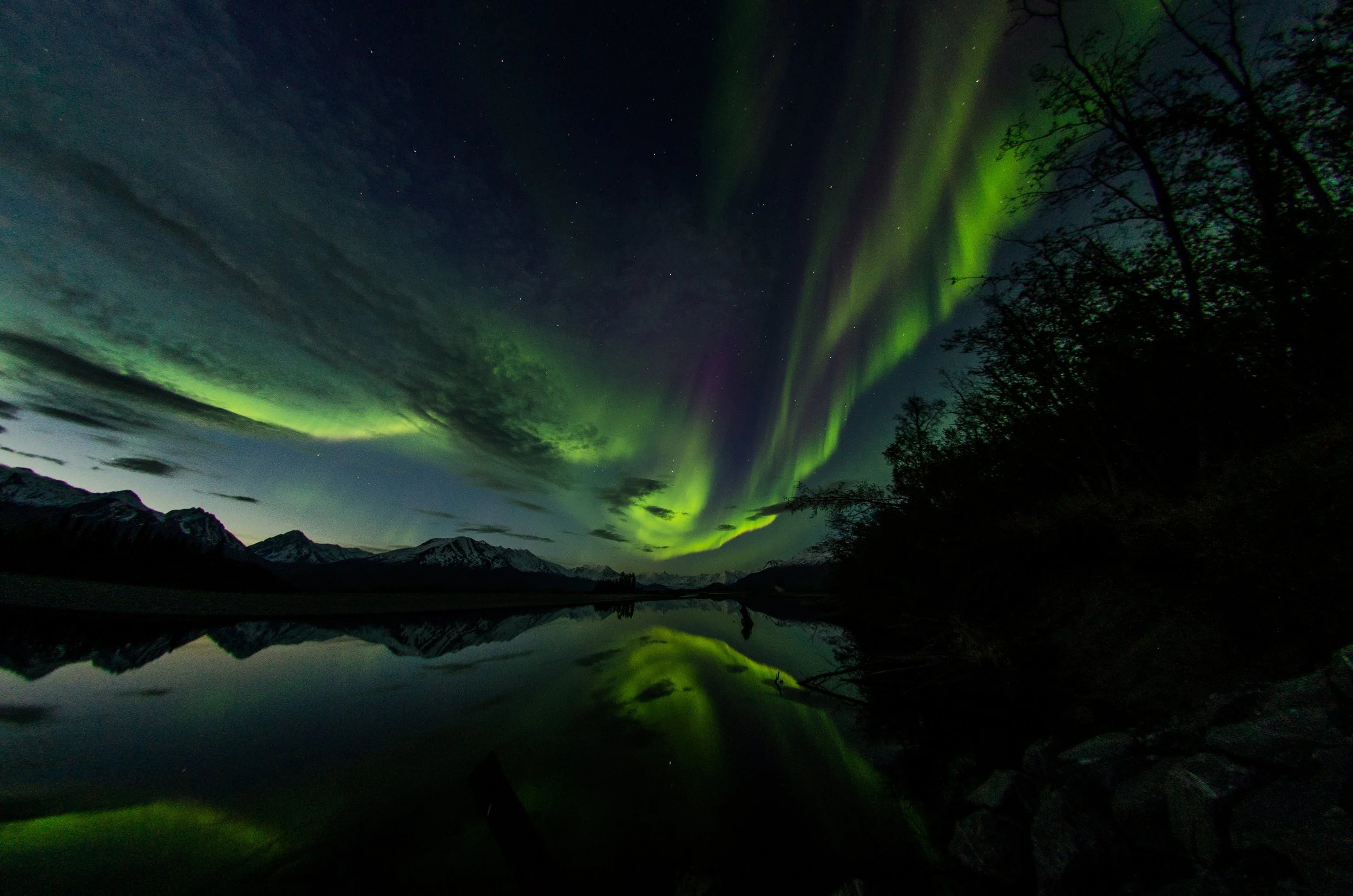 Northern lights (aurora borealis) over a mountain landscape at night, with a lake reflecting the sky and sparse trees on the right.