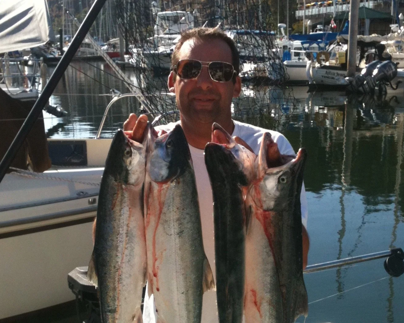 Man on a dock holding two large freshly caught fish, with boats and marina in the background.