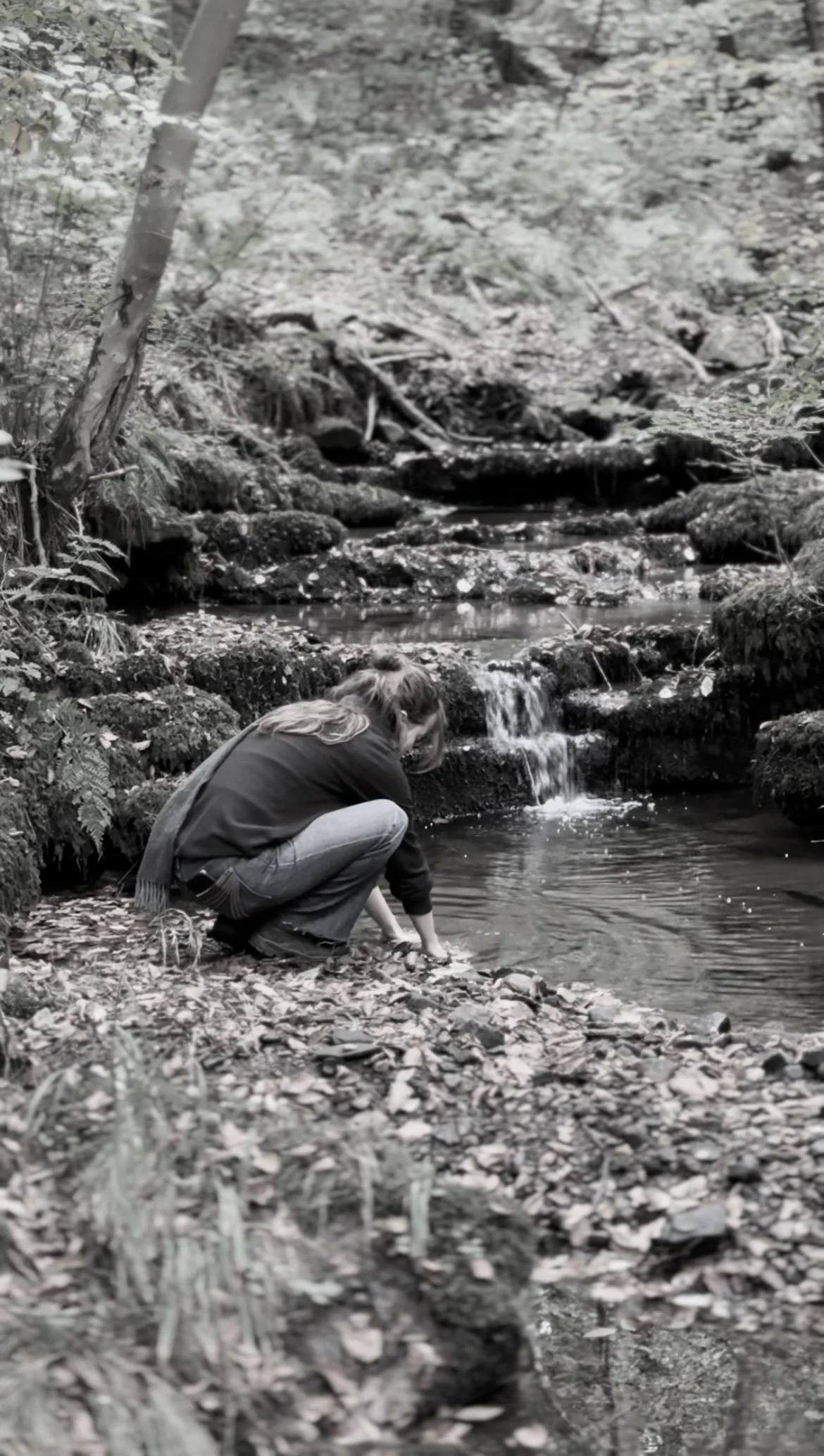 Olivia crouching by a small creek in a forested area of Northumberland, reaching into the water with her hand.