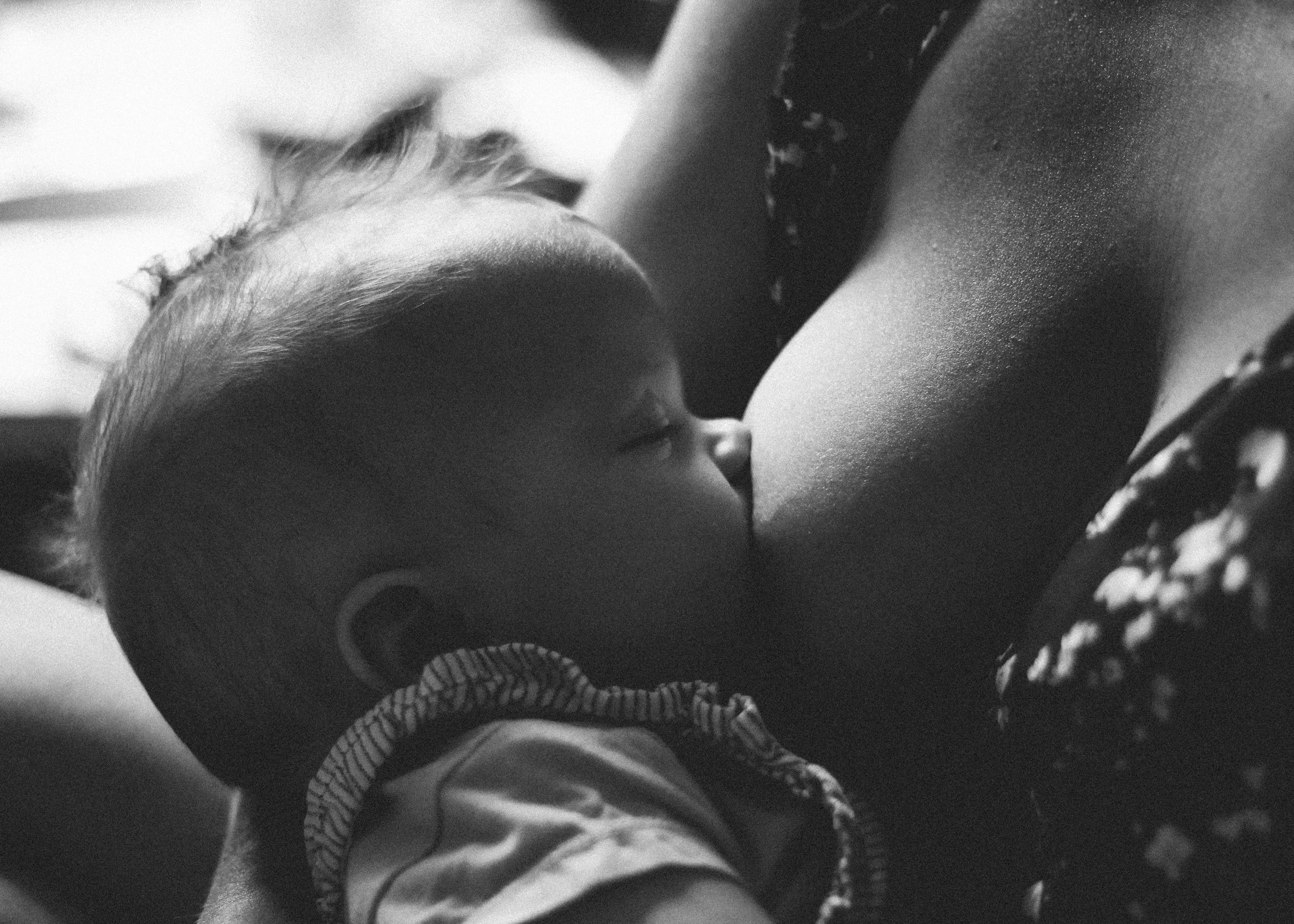 A close-up of a baby breastfeeding/chestfeeding, eyes closed and resting on an adult's chest in black and white.