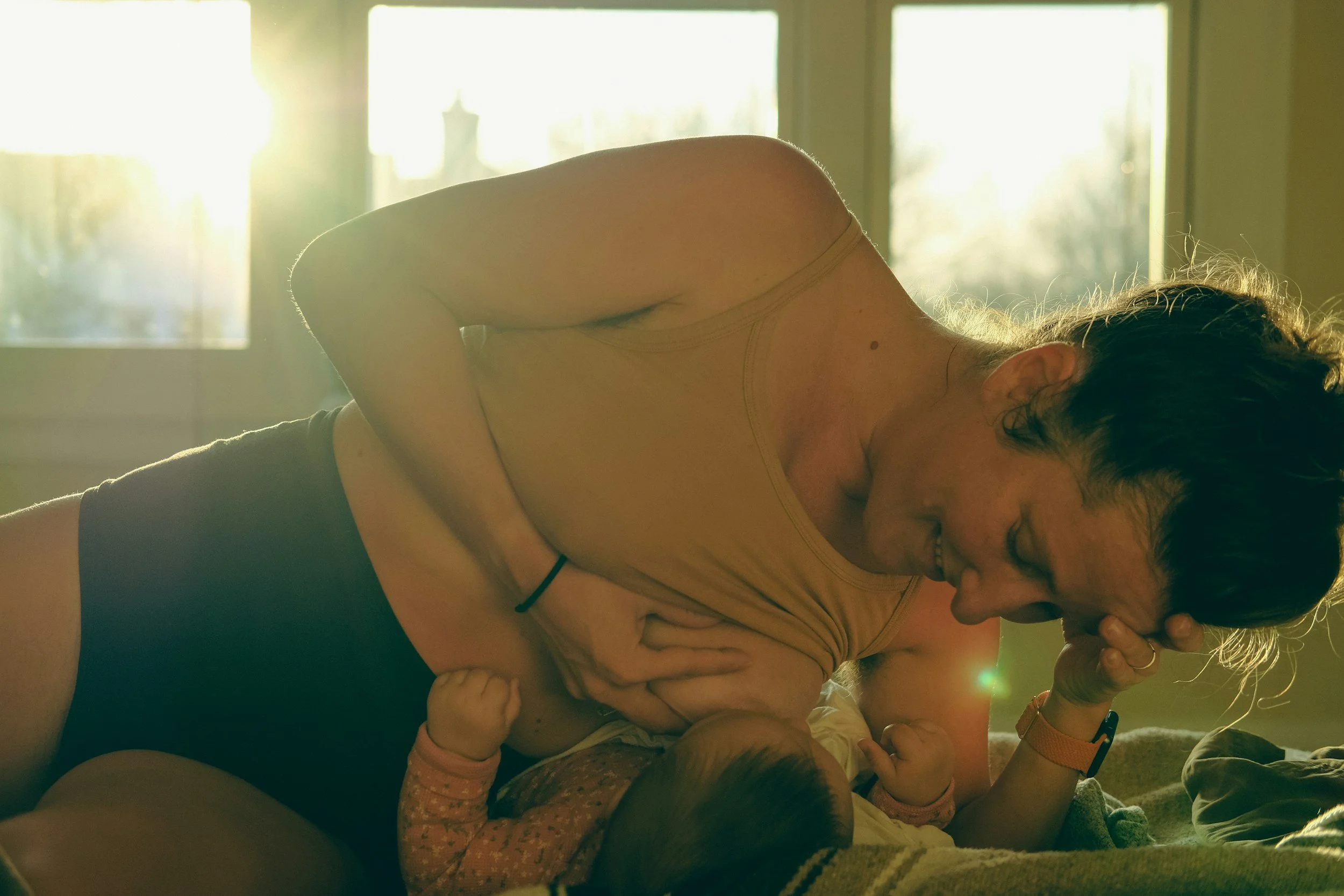A woman breastfeeding her baby in a side lying position. The mother is smiling down at her infant as sunlight gently shines in through the window.