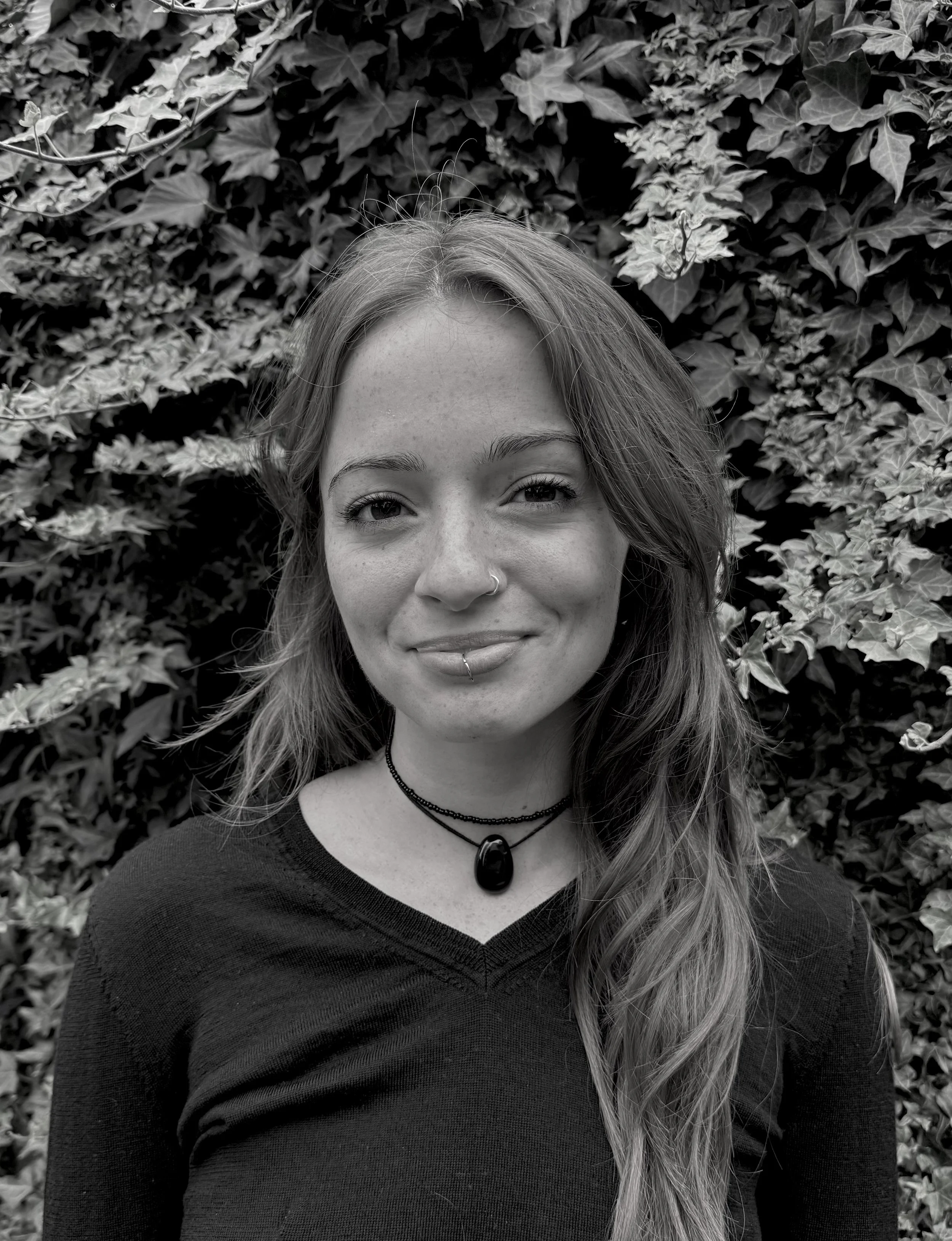Black and white headshot of Olivia, a breastfeeding consultant and doula. She has long wavy hair, smiling, wearing a nose ring and lip piercing, layered necklaces, and a black top, standing in front of dense ivy leaves.