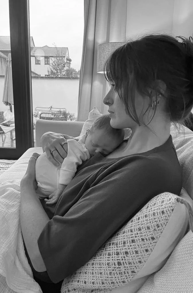 Olivia, doula and breastfeeding consultant, is sitting on a sofa with a newborn baby held close to her chest. A window in the background shows a residential backyard.