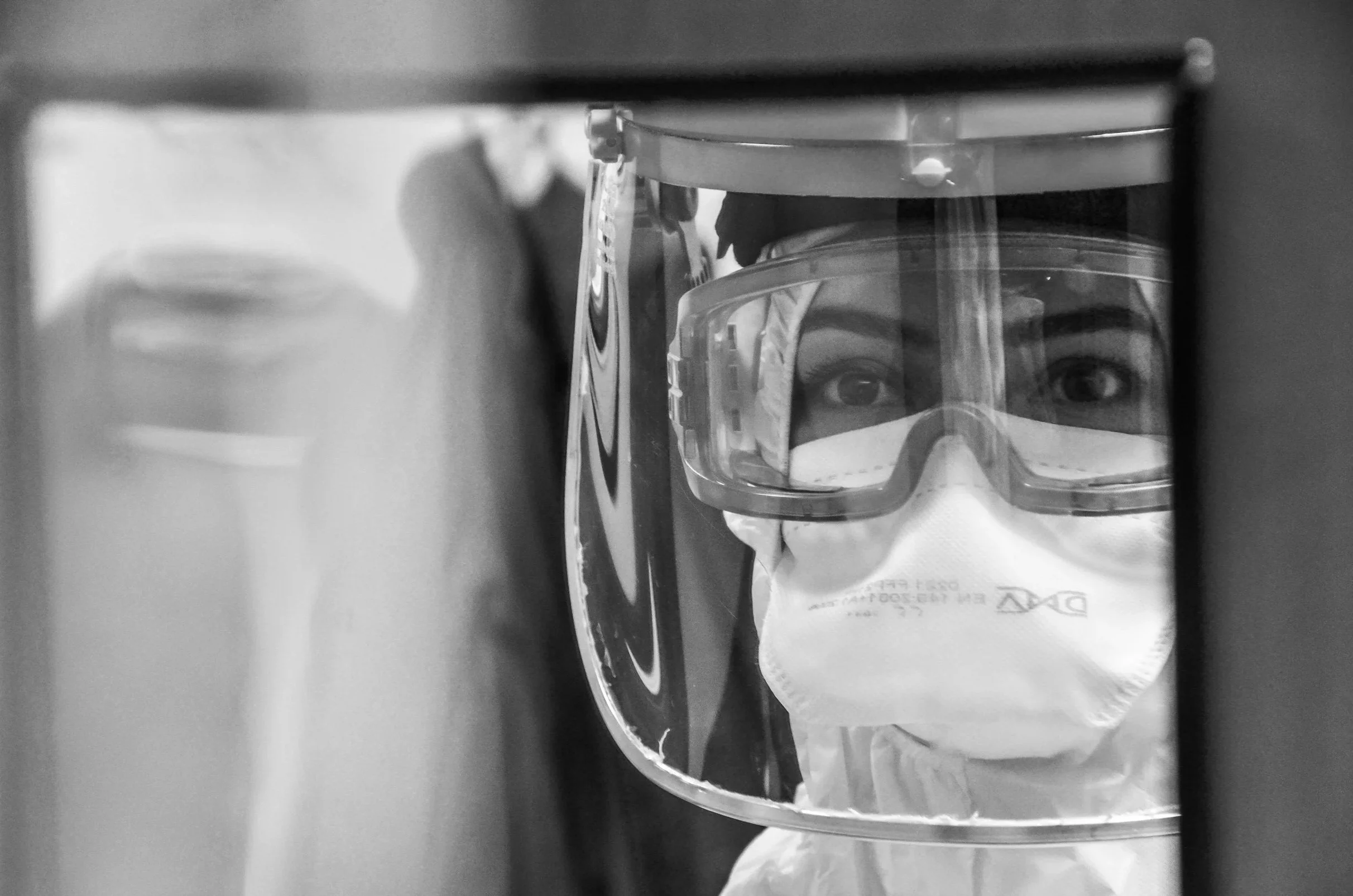 A person wearing a face mask, protective goggles, and a plastic cover looking into a mirror.