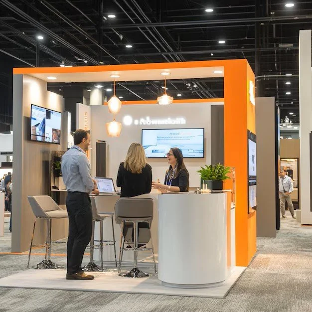 Trade show booth with three people conversing, two women and one man, surrounded by display screens, a white curved counter with a potted plant, and orange and gray accents.