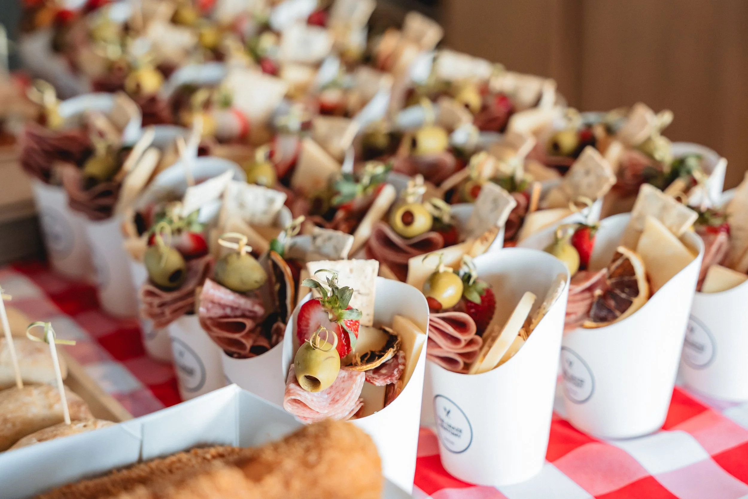 Small sandwiches with slices of meat, cheese, strawberries, and green olives served in white cups on a red and white checkered tablecloth.