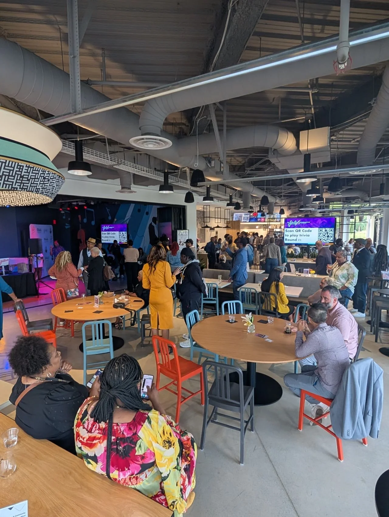 A lively indoor event space or restaurant with people gathered around tables, some engaging with their phones. There are large screens displaying QR code instructions for playing trivia, and a stage area with musical instruments and lighting. The ceiling is industrial with exposed ductwork and pipes.