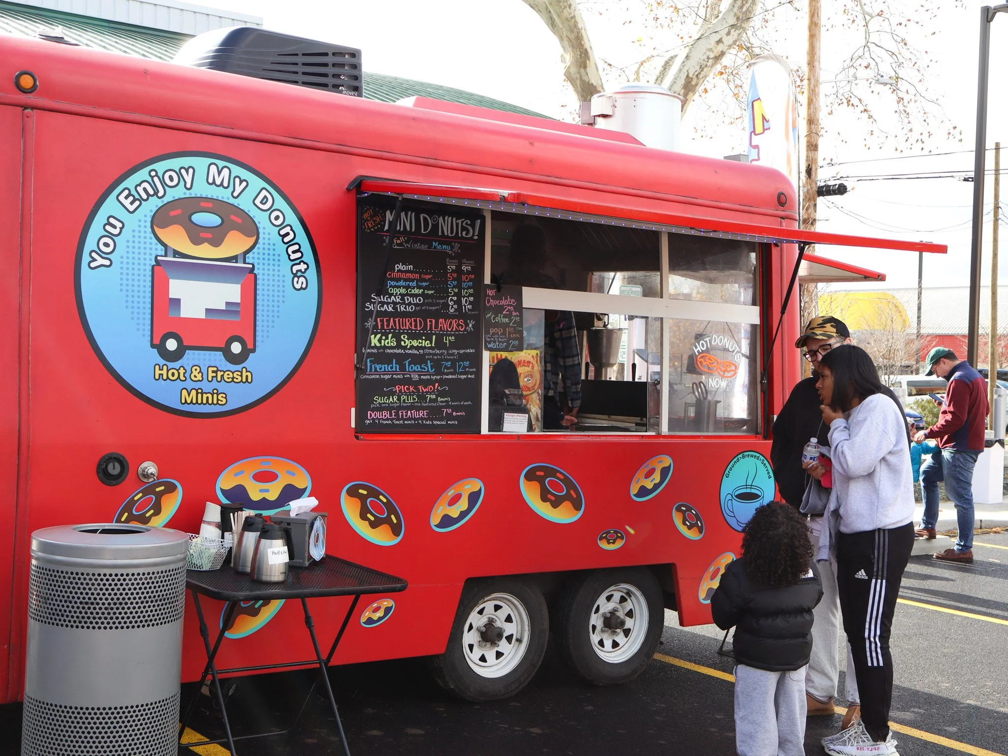 Red food truck with donut logo and sign that reads "You Enjoy My Donuts" and "Hot & Fresh Minis." Customers, including a woman and a child, stand in line at the truck, which has a blackboard menu with various donut flavors and prices, and several decorative donut stickers on its side.