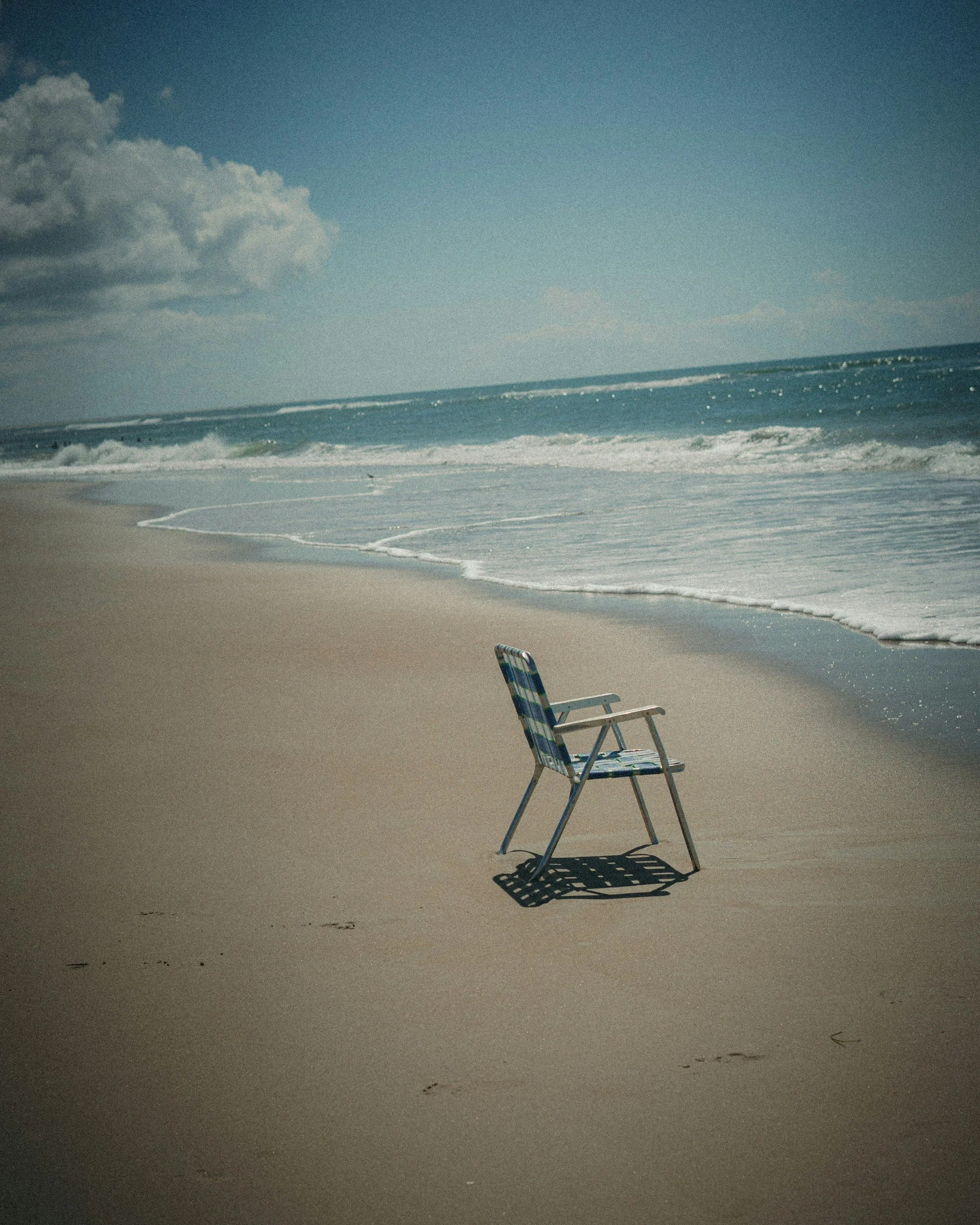 Empty plaid beach chair on sandy shore near ocean waves, with cloudy sky overhead.