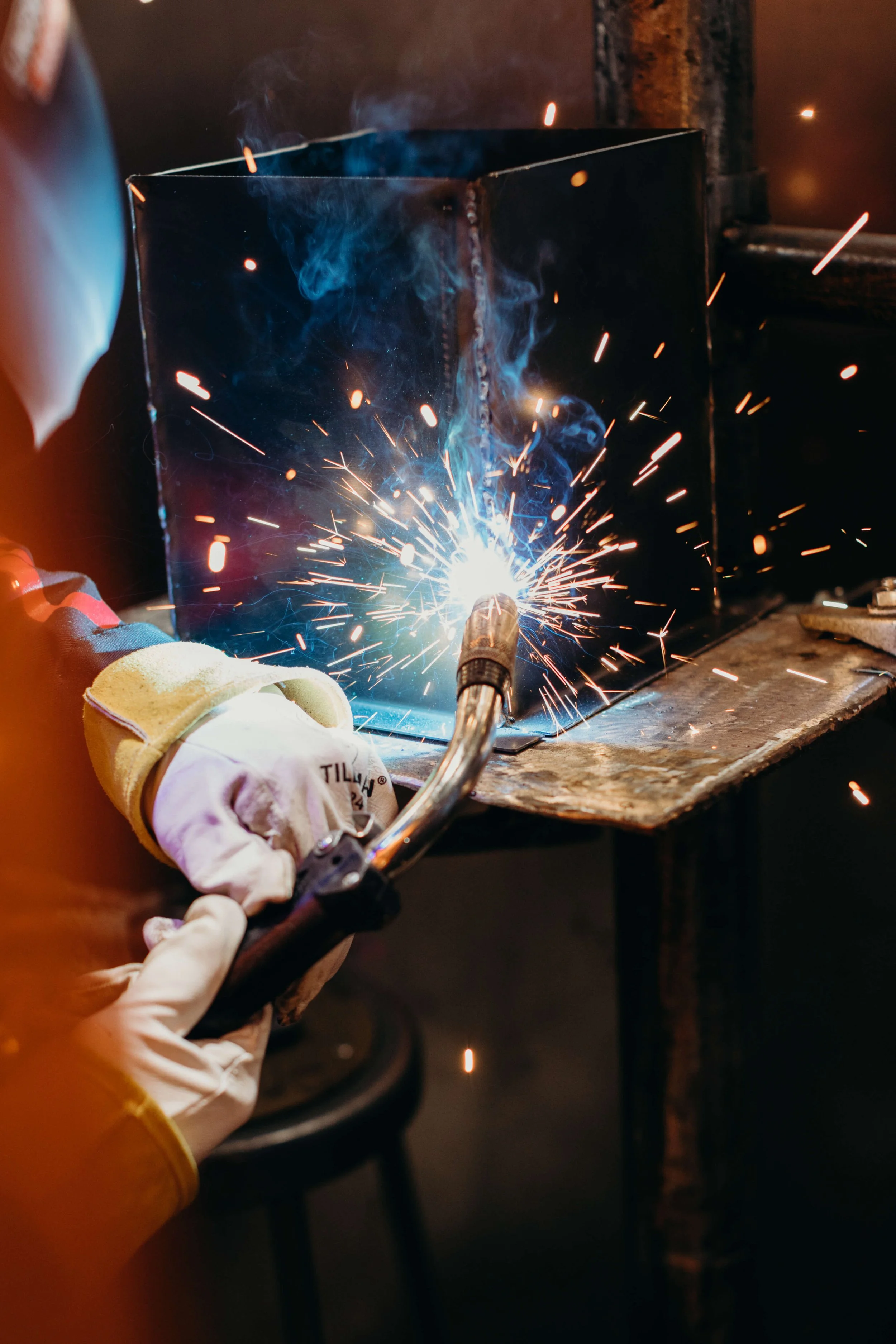 Welding metal piece with an arc welder, sparks flying, person wearing protective gloves and face shield, workbench and tools nearby.