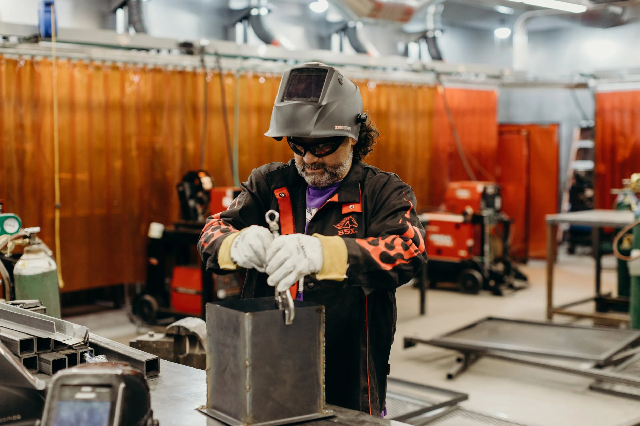 A man wearing welding protective gear, including a helmet and gloves, working with metal pieces in a welding shop with orange safety curtains in the background.