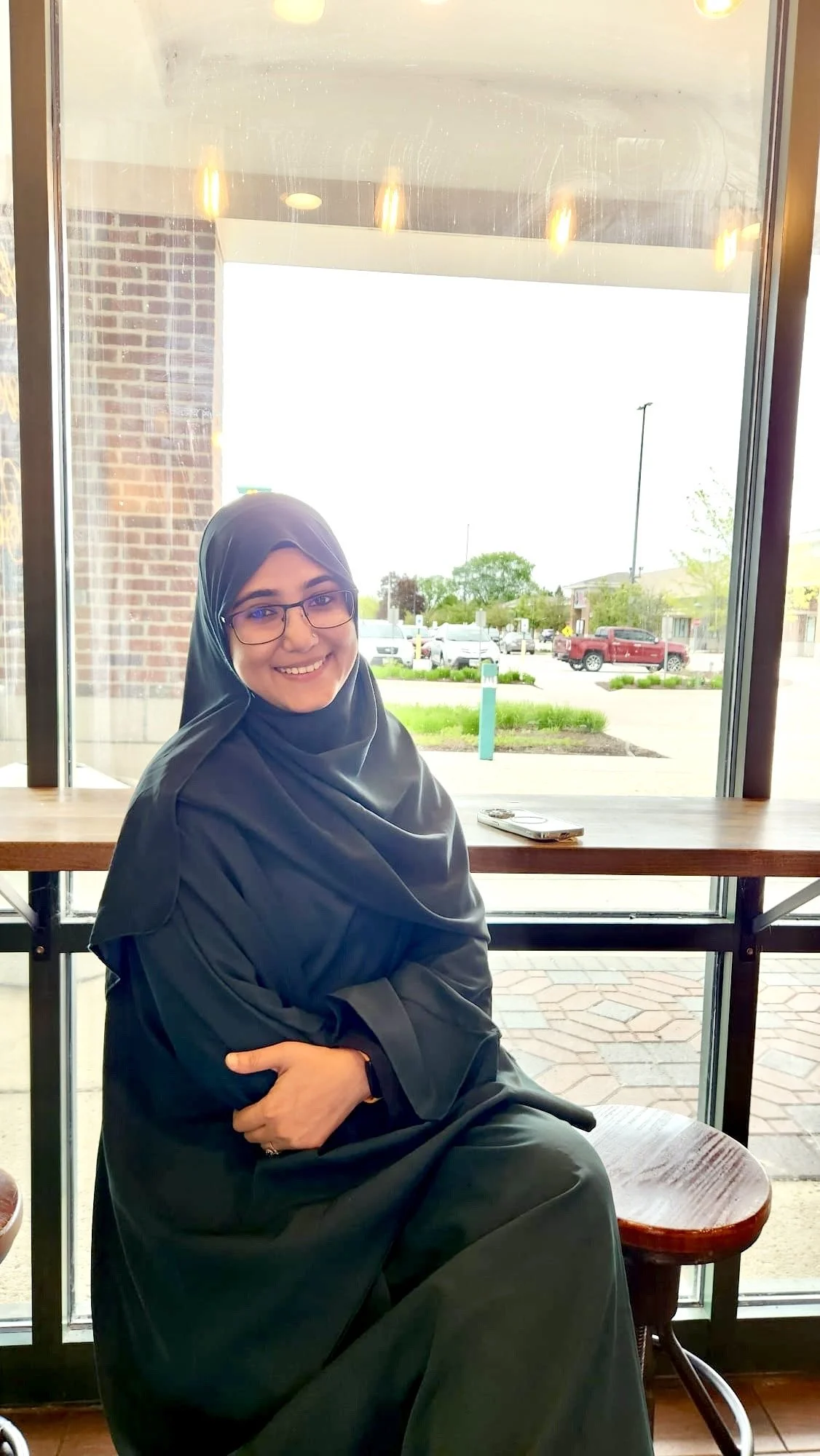 A woman wearing glasses and a dark headscarf, smiling while sitting by a large window in a cafe or restaurant. Outside the window, there is a parking lot with cars and trees.