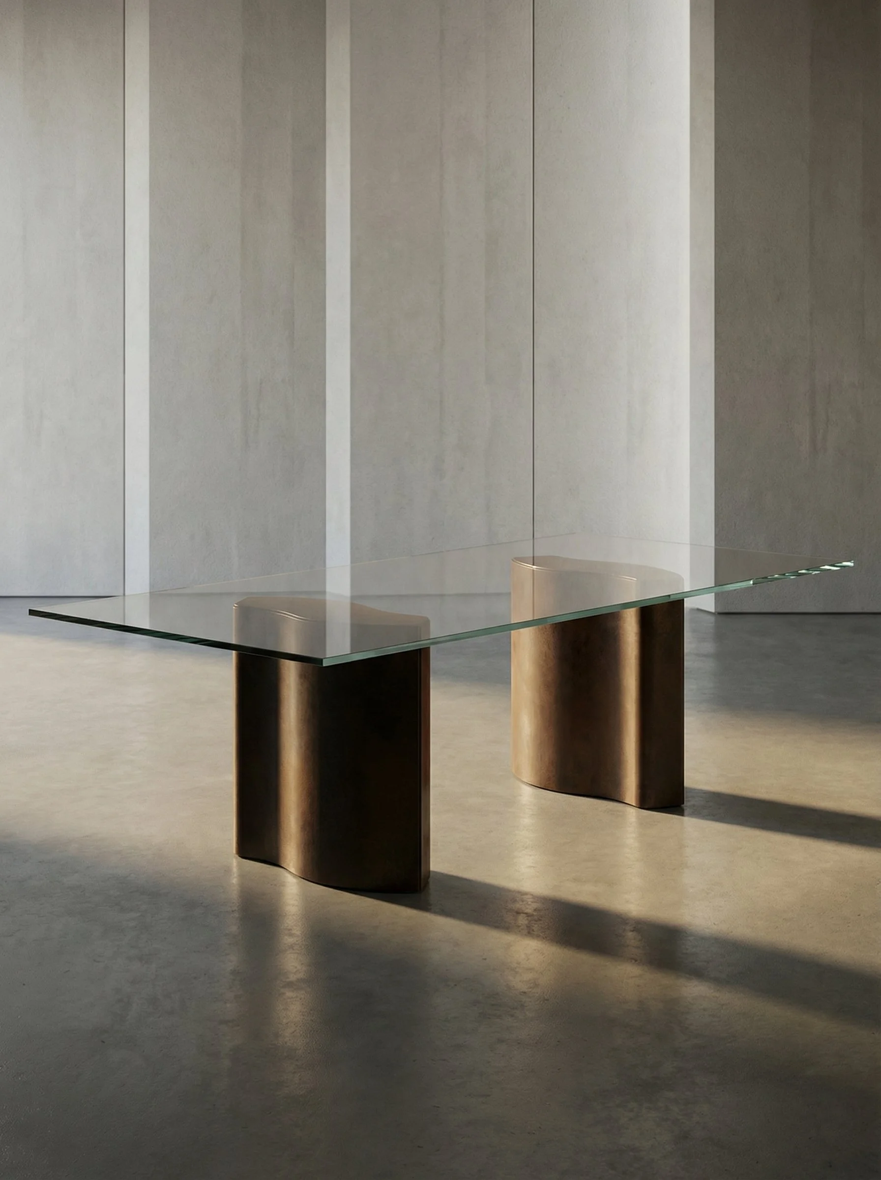 Tavolo con piano in vetro e gambe in metallo con finitura dorata, in uno spazio con pareti di colore chiaro e pavimento in cemento.