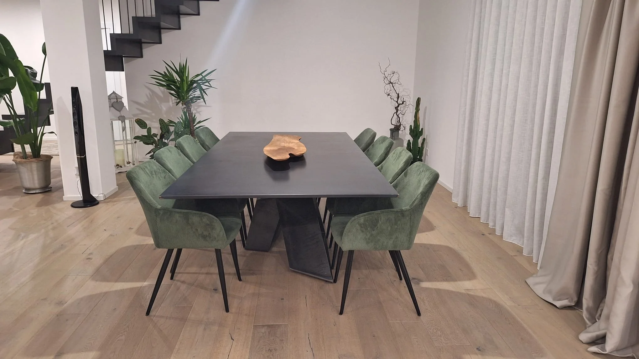 Sala da pranzo moderna con un grande tavolo nero e sedie verdi in velluto, decorata con piante verdi e un ramo secco in un vaso sulla parete.