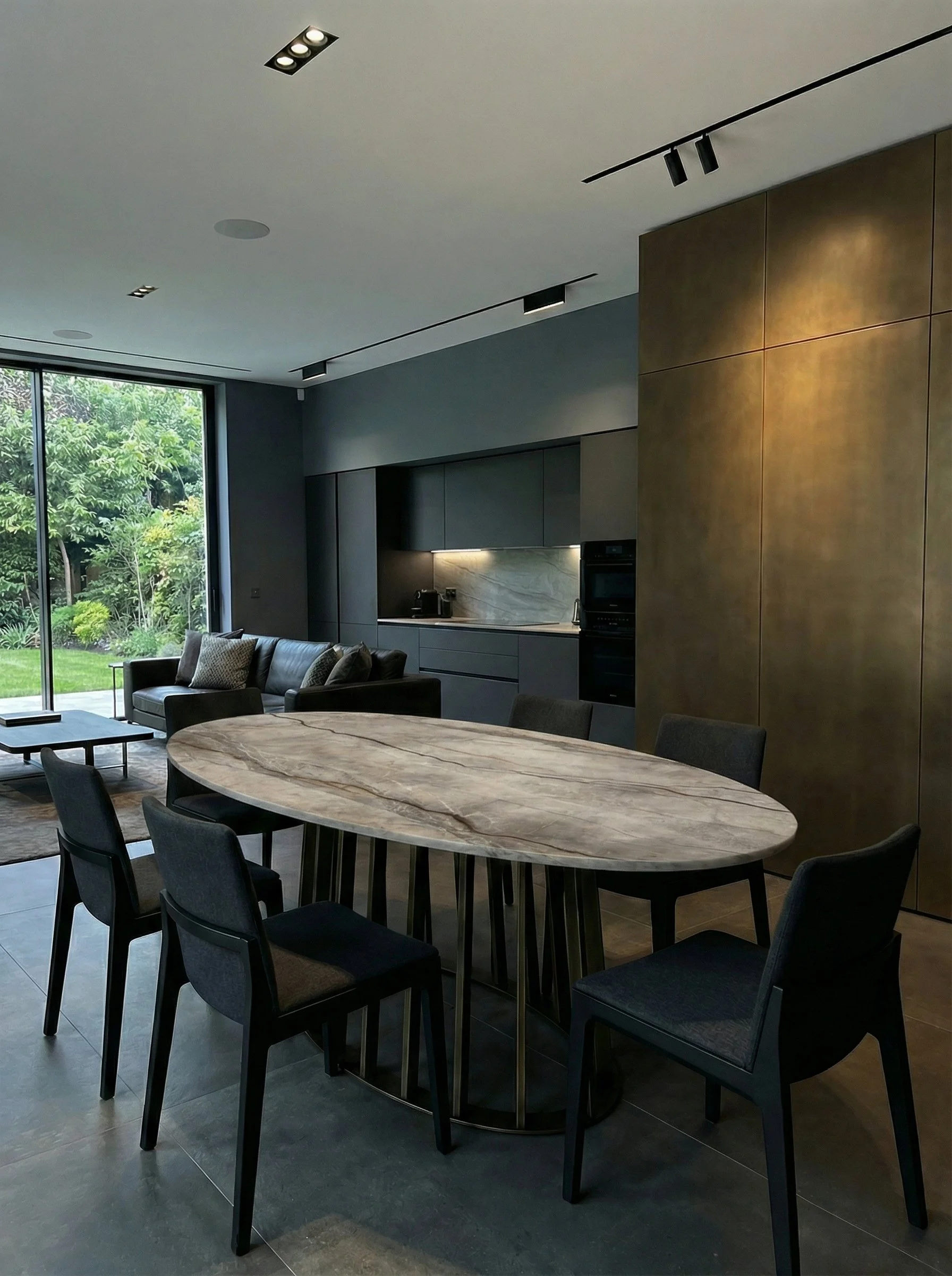 Interior di una cucina e sala da pranzo moderna con tavolo rotondo in marmo, sedie nere, zona soggiorno con divano e grandi finestre con vista su un giardino verde.