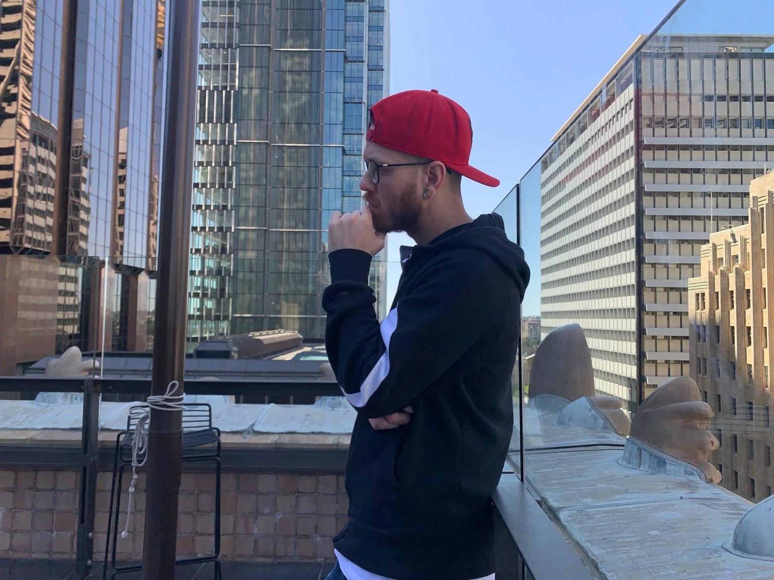 Intalekz in a red cap and dark hoodie beside a rooftop glass railing and city buildings.