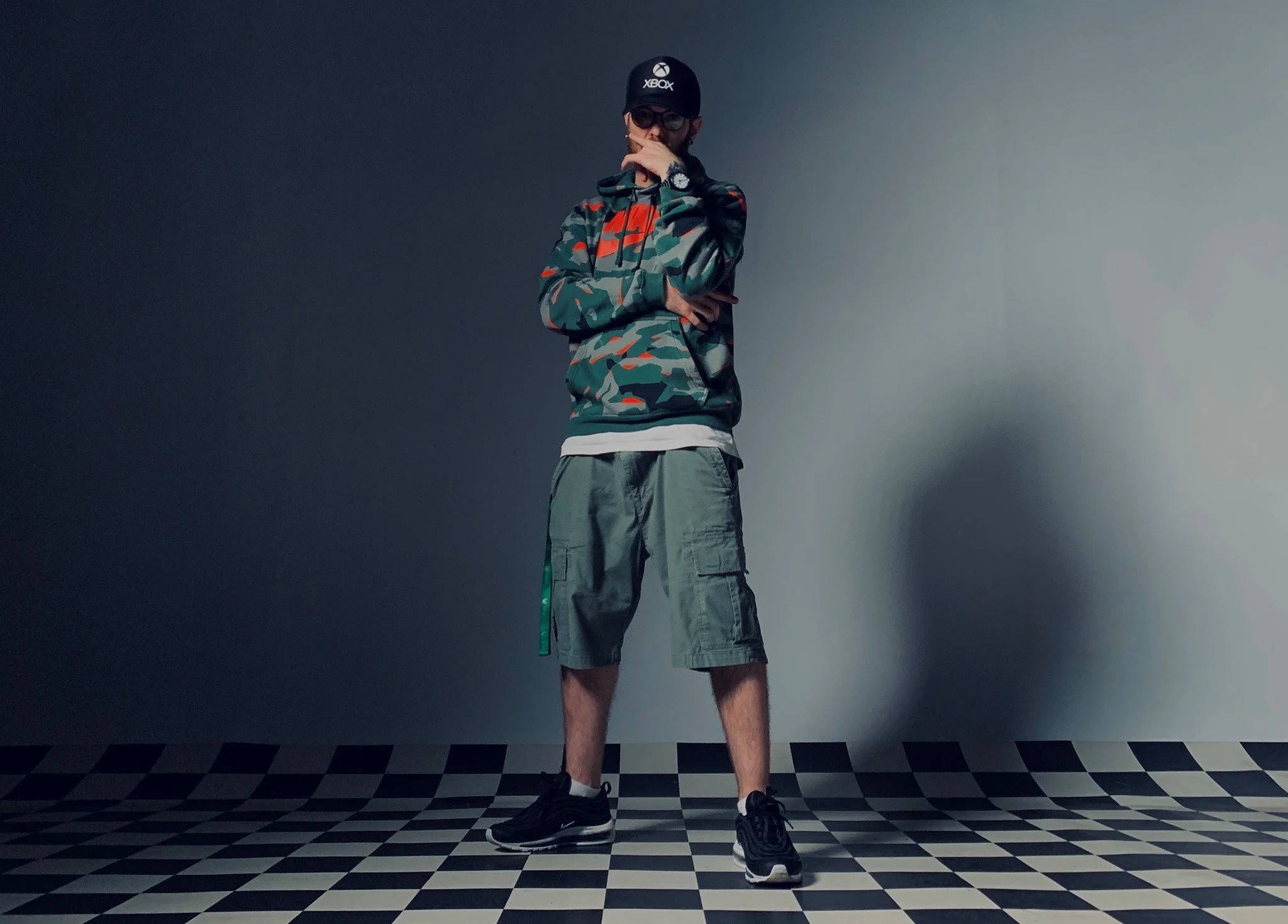 Full-length portrait in a camouflage hoodie and cap against a gray backdrop.
