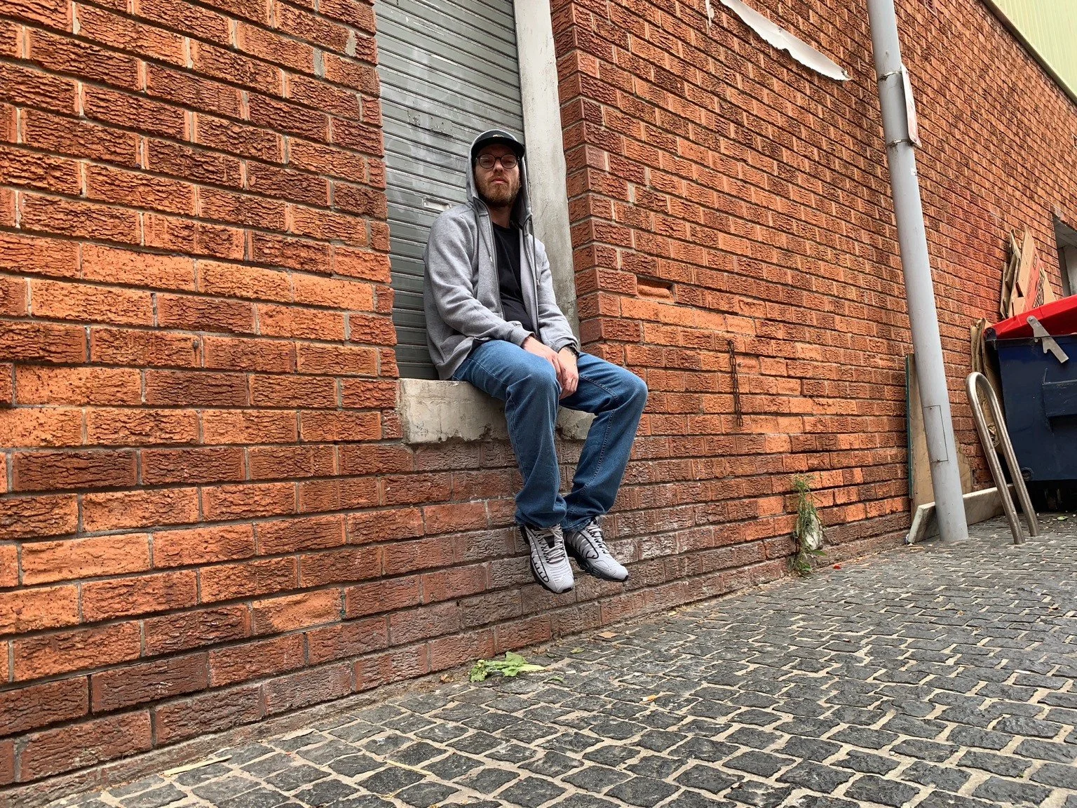Intalekz in a grey hoodie and jeans seated beside a brick street wall.