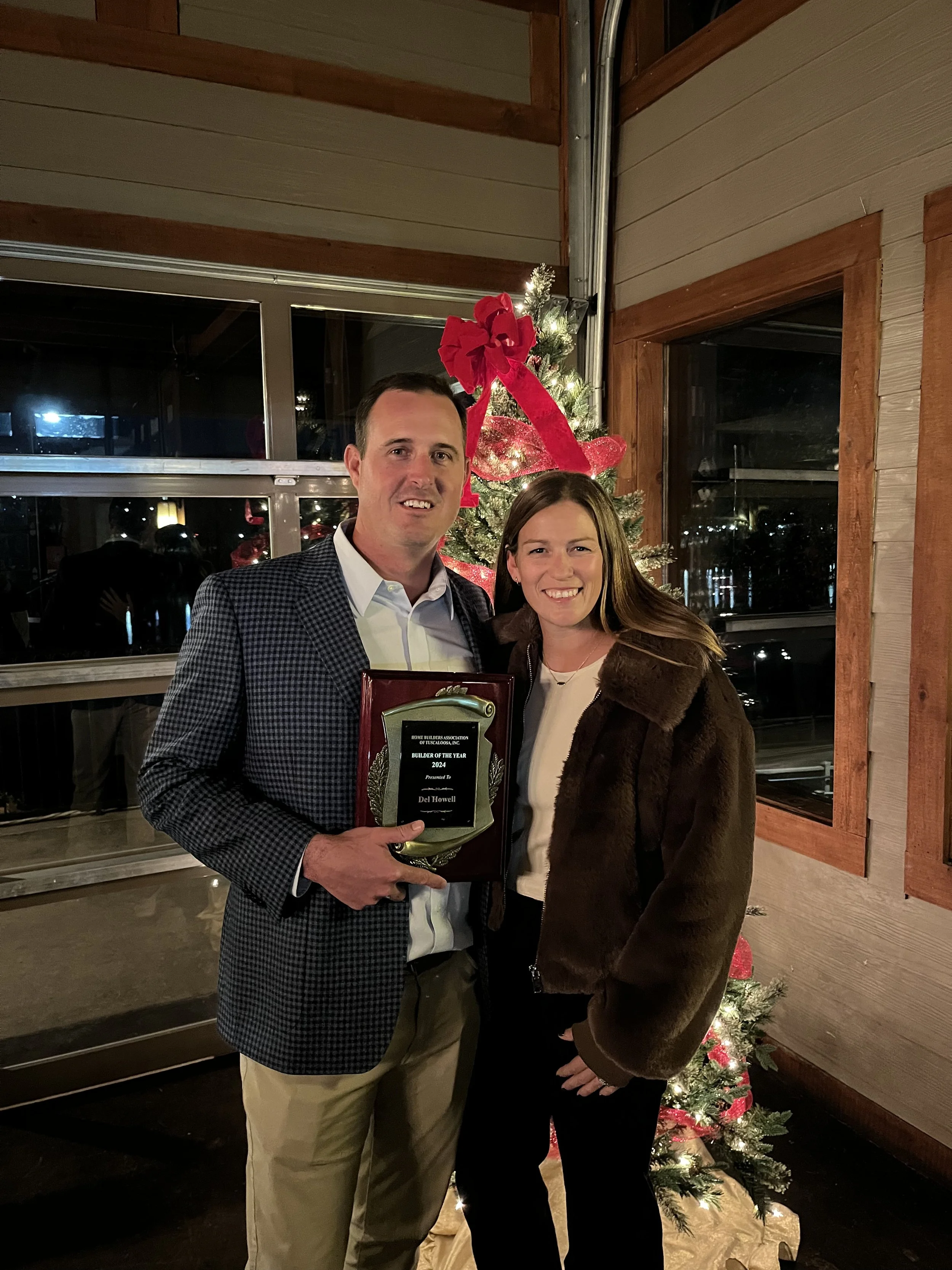 A man and woman standing together at Christmas, holding an award plaque, in front of a decorated Christmas tree with red ribbon and bow. They are indoors with wooden and glass window elements.