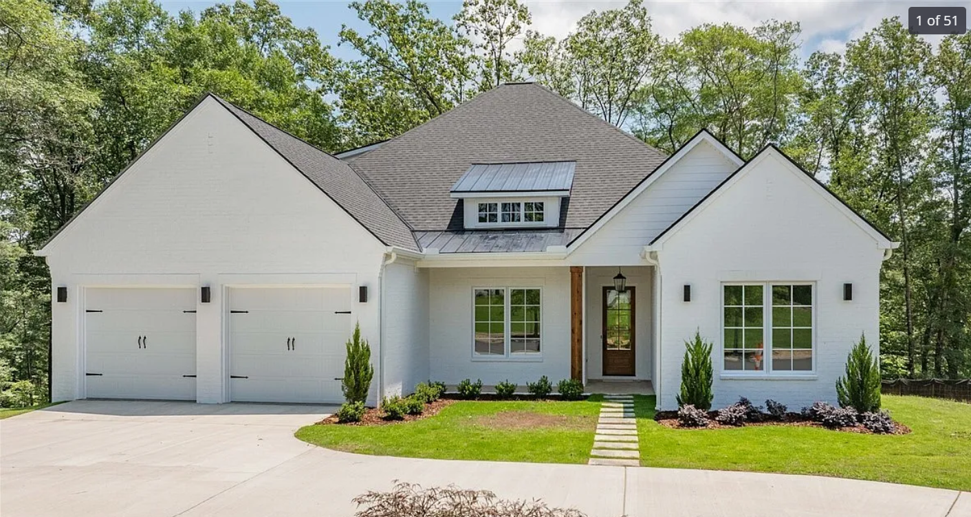 Modern white house with a gray roof, double garage, front door, large windows, small landscaped yard, and trees in the background.