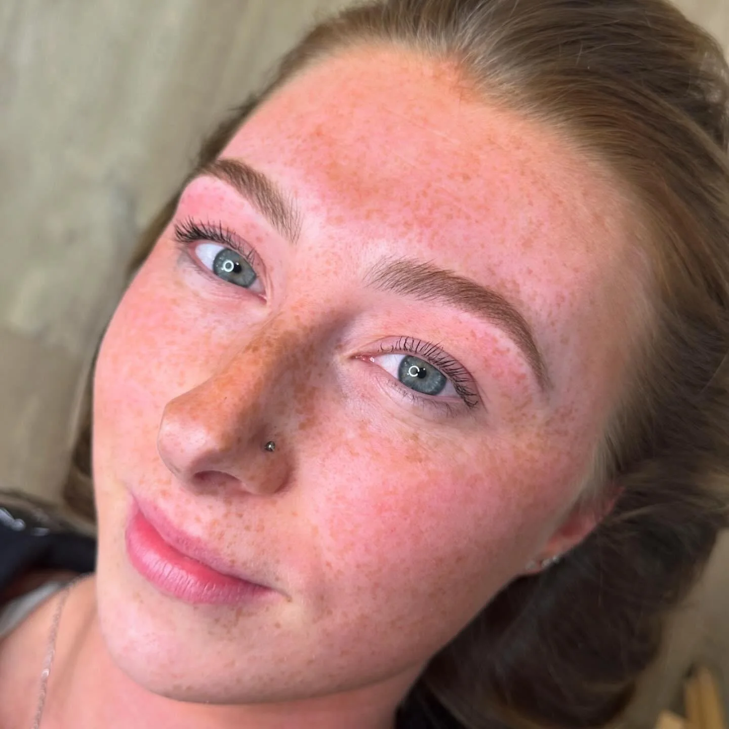 Close-up of a young woman with light skin, freckles, blue eyes, and a nose piercing, lying down and looking at the camera.