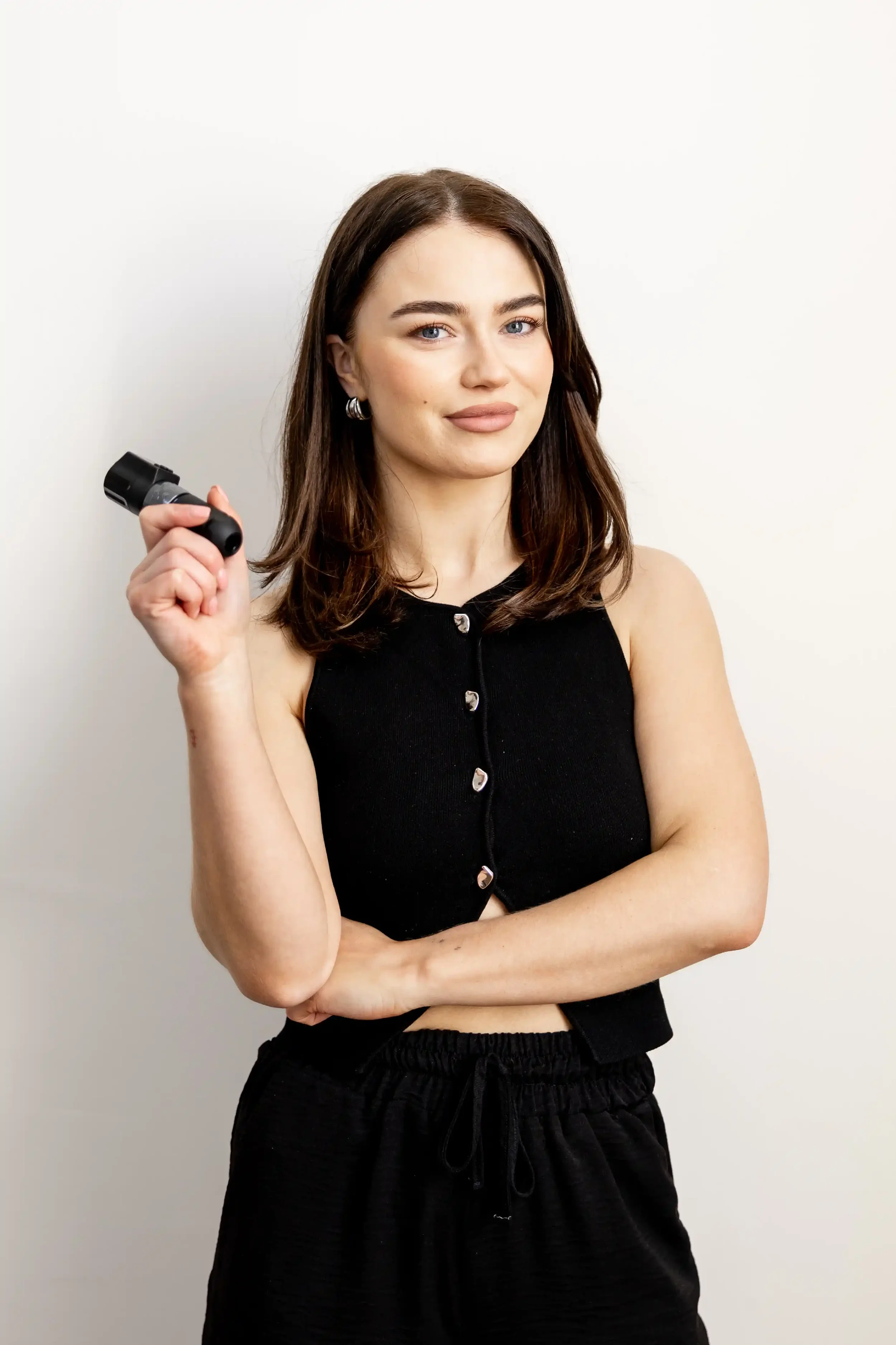 Evan, wearing a black sleeveless top and black drawstring pants, holding a black dumbbell in her right hand, standing against a plain white background.