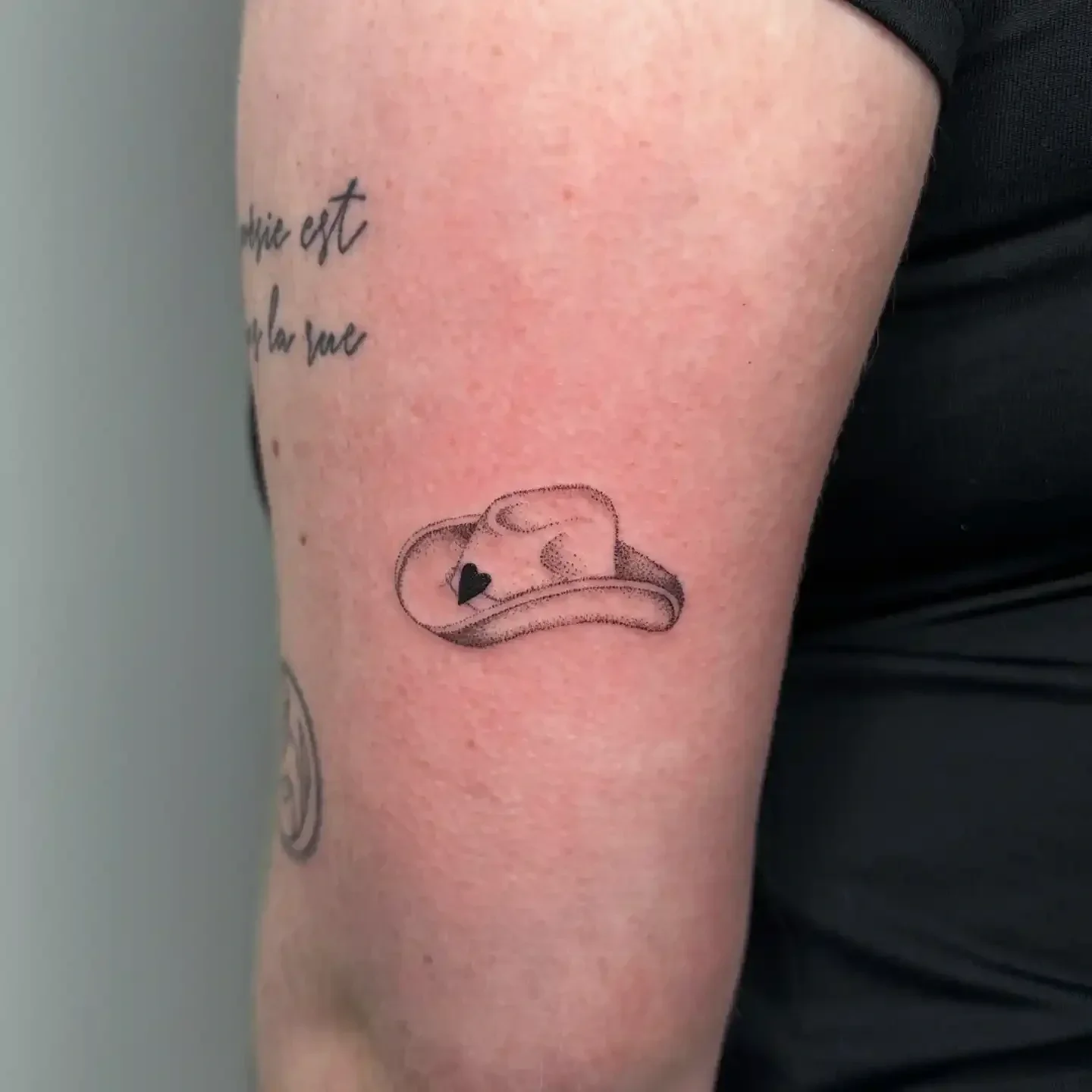 Tattoo of a simple outline of a cowboy hat with a love heart on, on a person's arm.
