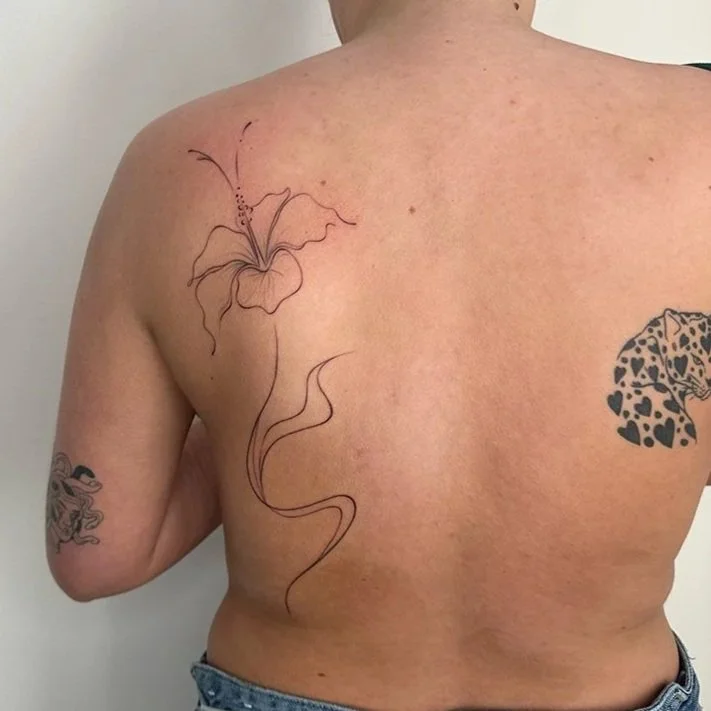 A person's back with two tattoos: a delicate, outlined flower with flowing lines near the shoulder and a black leopard pattern tattoo on the right arm.