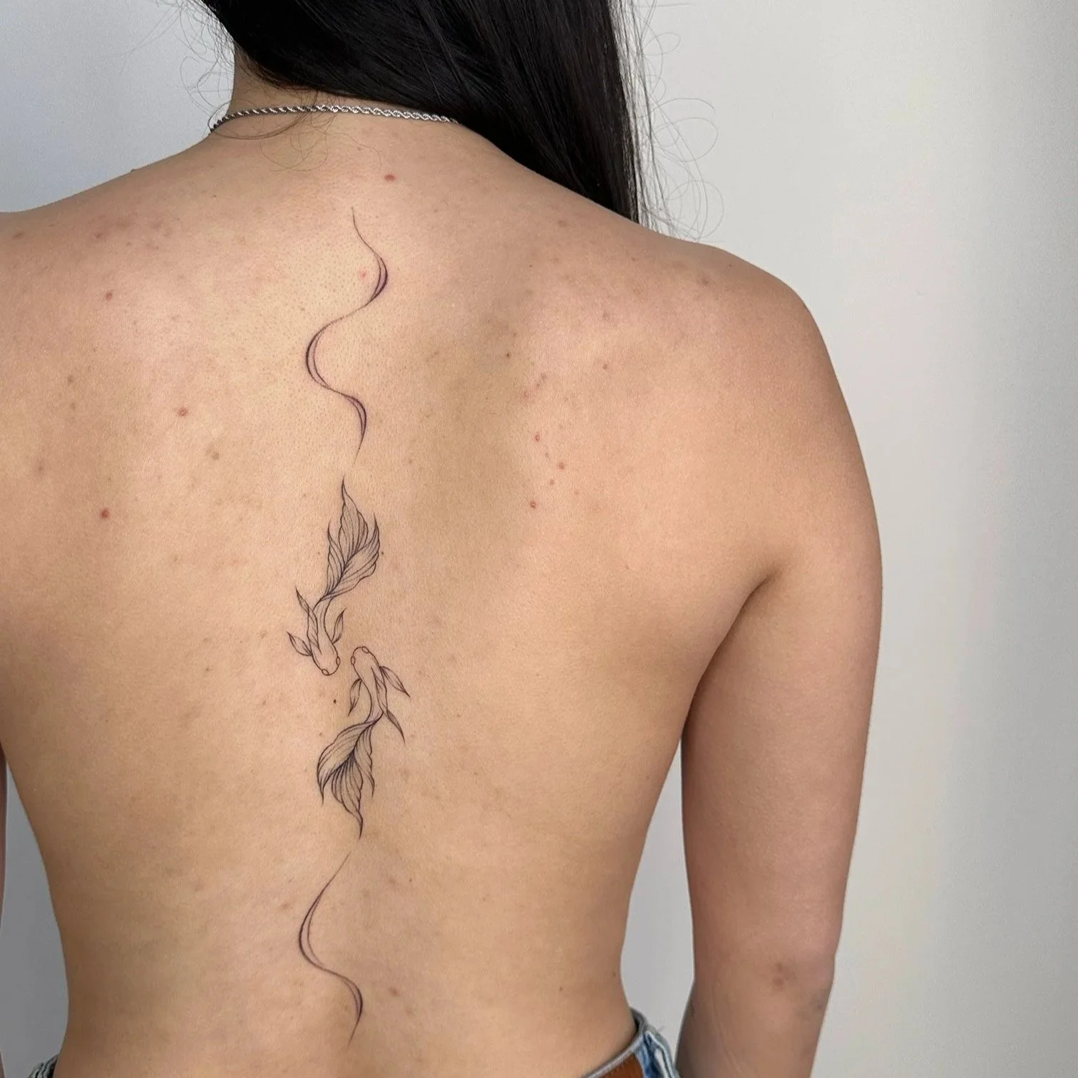 Back view of a woman with black hair, showing a vertical line art tattoo of two koi fish with flowing fins on her back.
