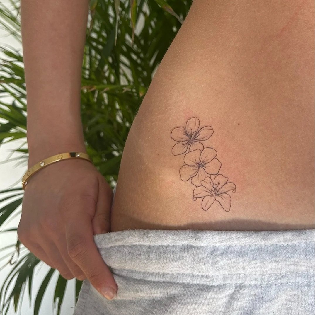 Line art tattoo of three hibiscus flowers on a person's side, with a person holding their waistband with one hand and wearing a gold bracelet.