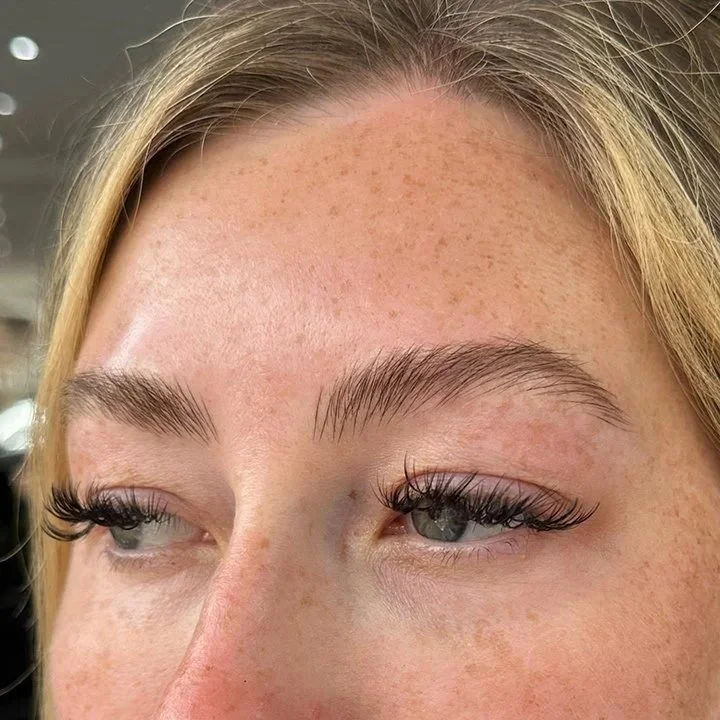 Close-up of a woman's forehead, eyebrows, and eyes with eyelash extensions and natural freckles on her skin.