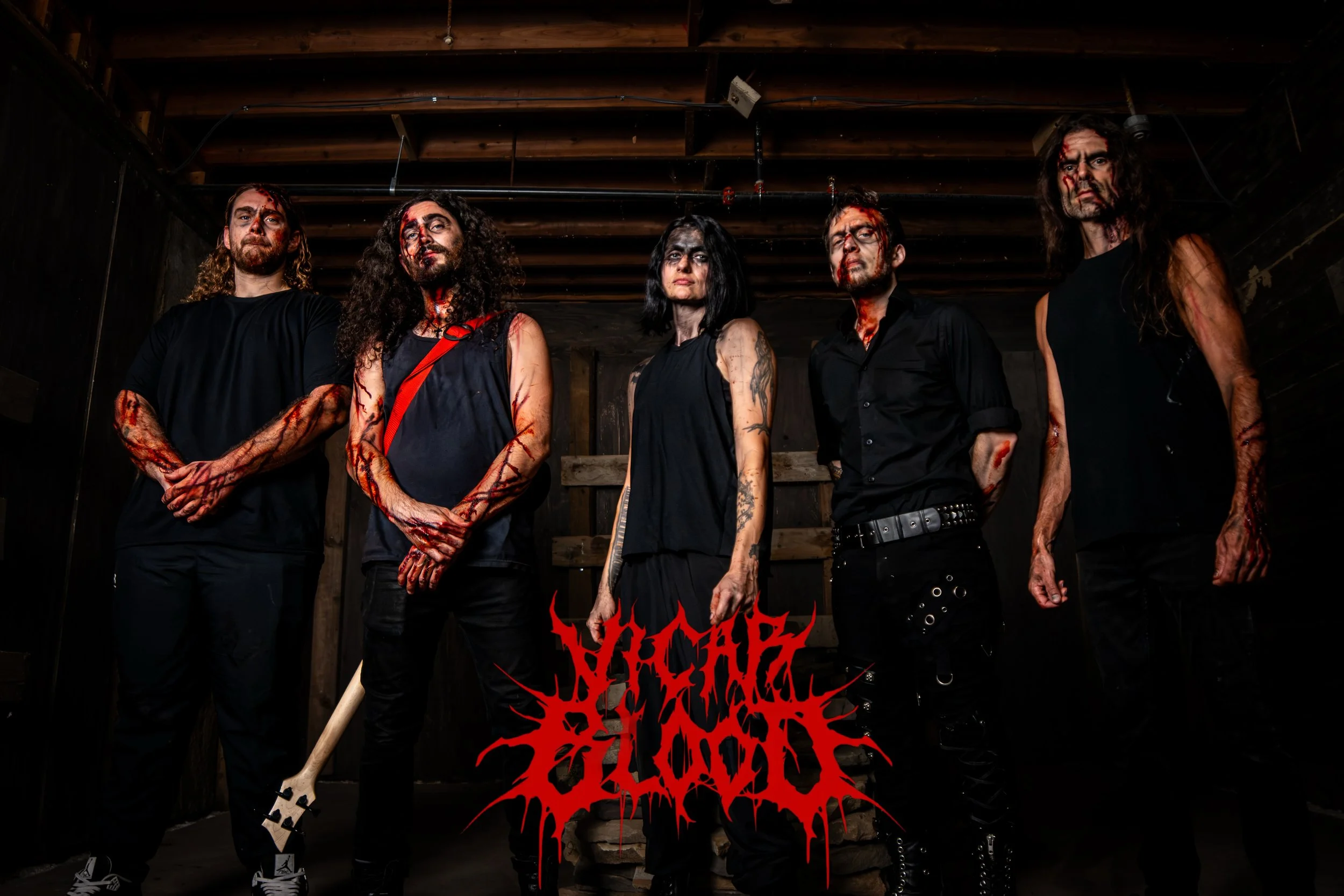A group of five people with dark makeup and fake blood, standing in a dark, wooden setting. They are dressed in black clothing, and there is red and black text overlay at the bottom of the image.