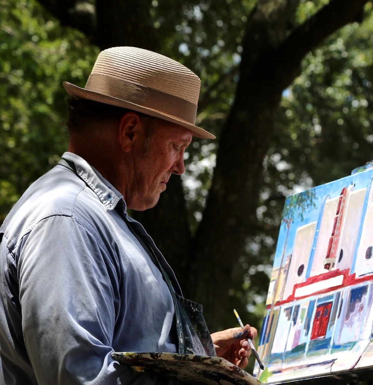 An elderly man wearing a straw hat and a light-colored shirt is painting on a canvas outdoors, with trees and greenery in the background.